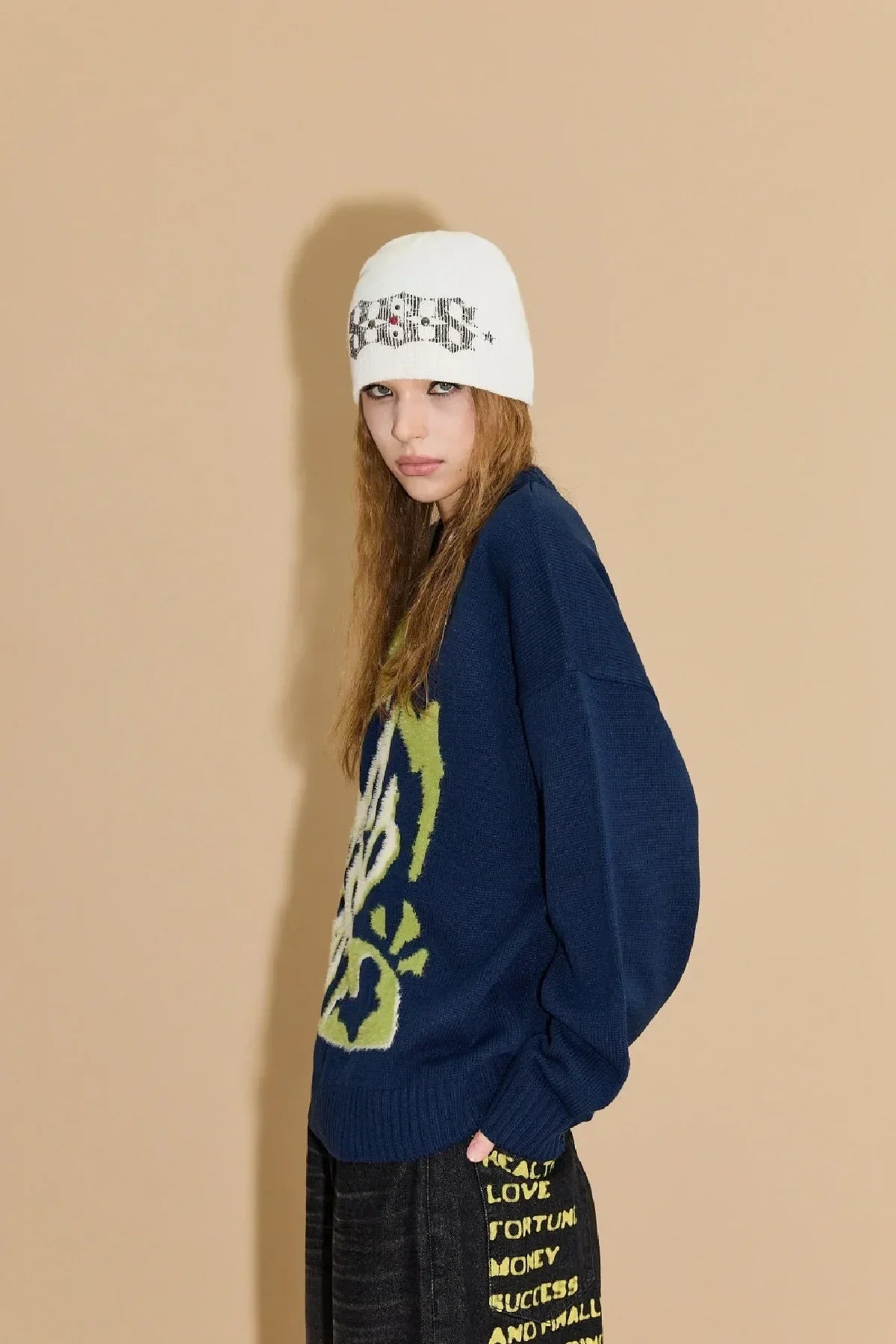 Model wearing white beanie with text and navy knit sweater with green graphic and black pants with yellow text