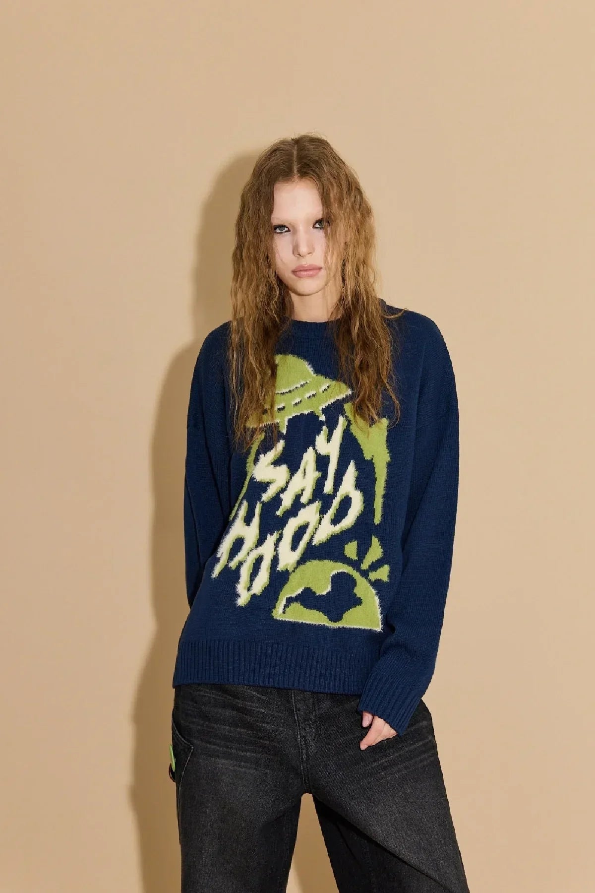 Woman wearing navy knit sweater with green UFO and 'Say Hood' graphic, posing against beige background