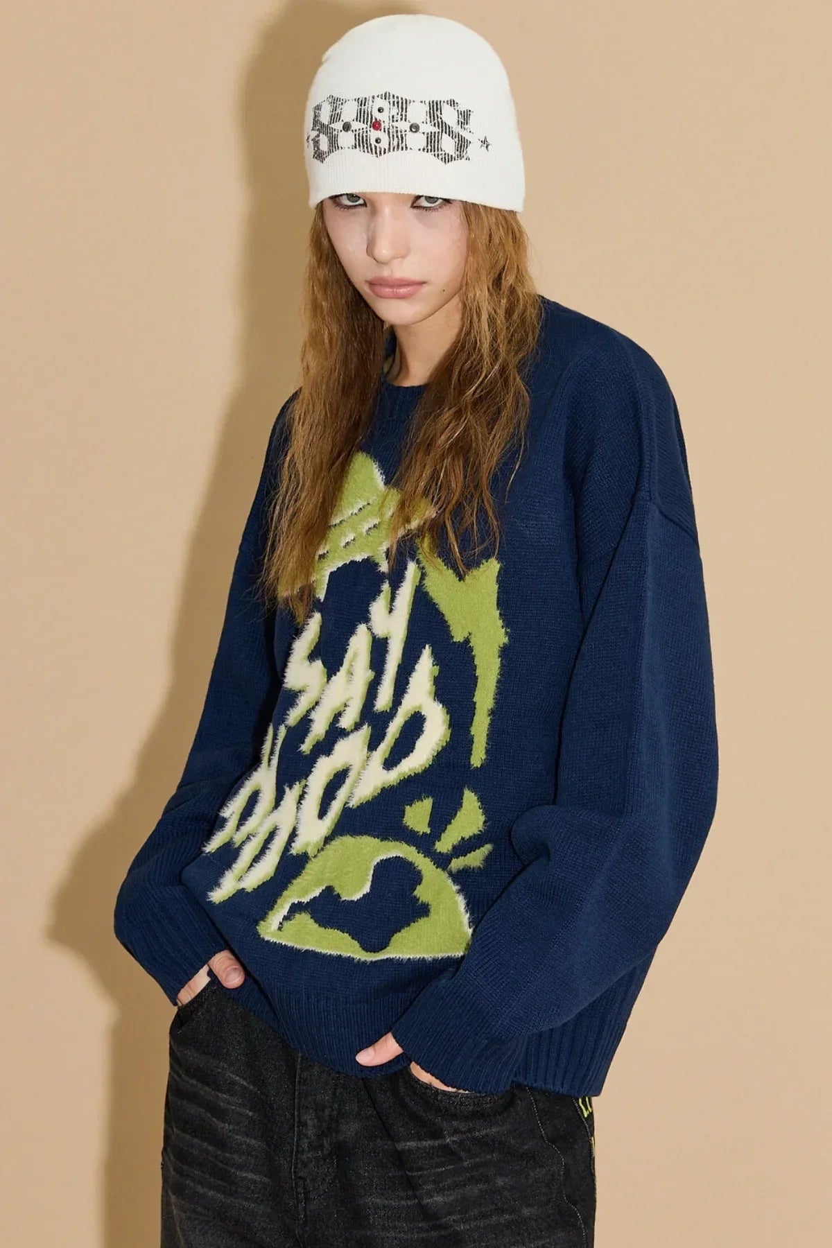 Model wearing navy knit sweater with green and white graphic text and white beanie hat in studio