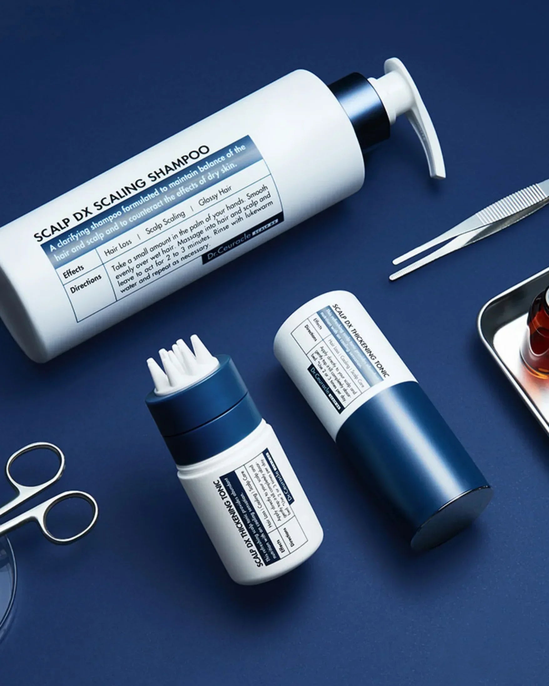 Scalp DX Scaling Shampoo and Thickening Tonic bottles with scalp massage tool and grooming scissors on blue background