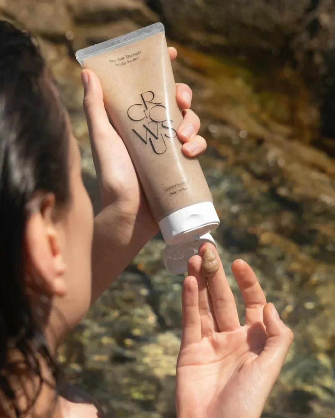 Person dispensing sea salt therapy scalp scaler exfoliating scrub from tube outdoors near water