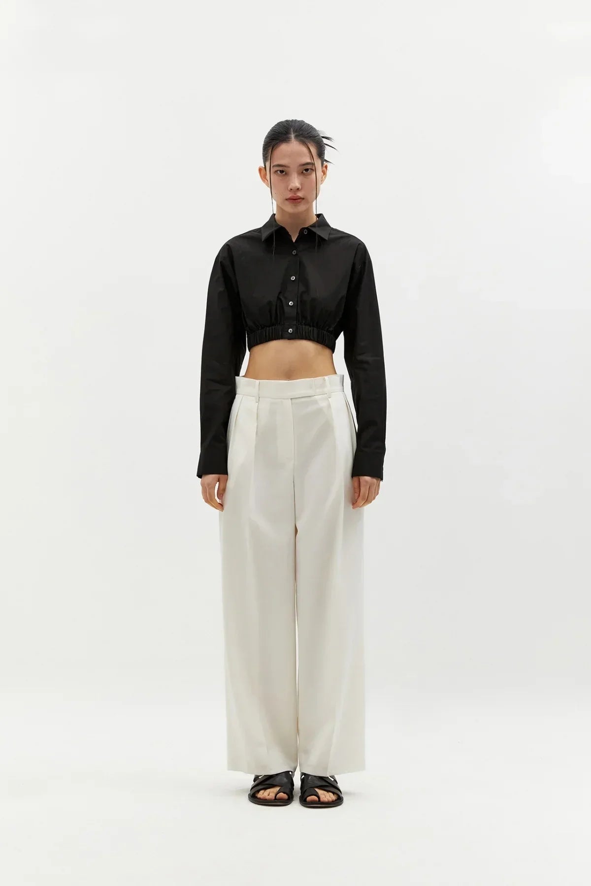 Model wearing black cropped cotton shirt with white wide-leg pants and black sandals on white background