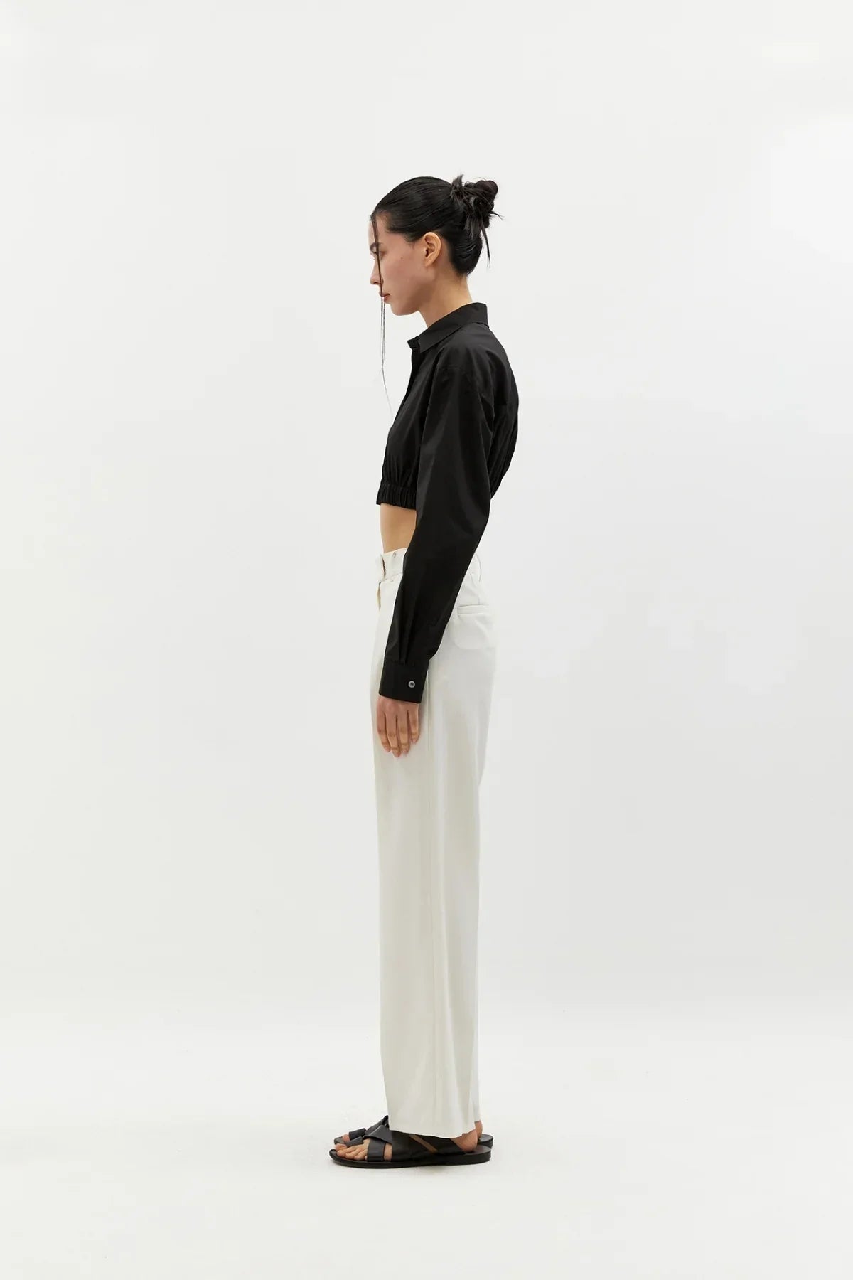 Side view of woman wearing black cropped shirt, white wide-leg pants, and black sandals on white background