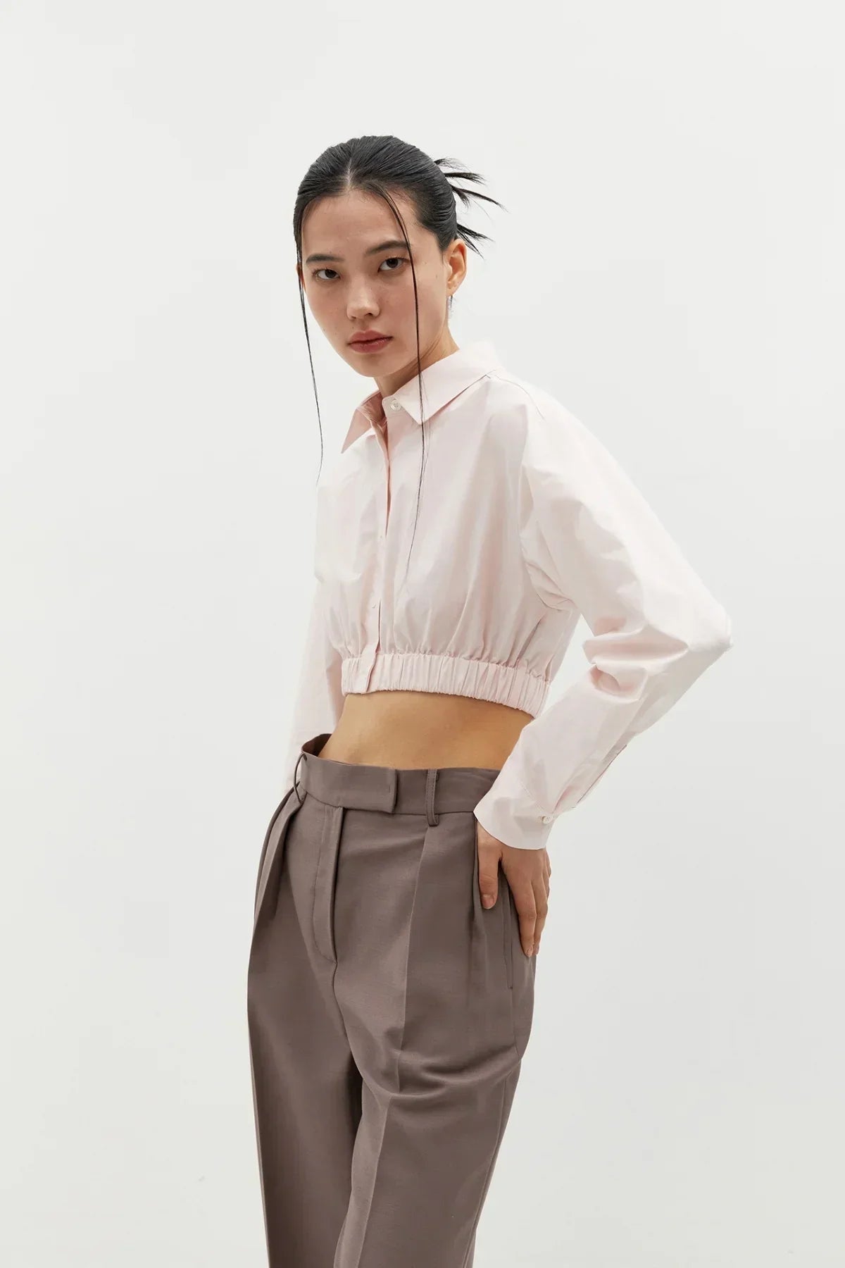 Model wearing light pink cropped cotton shirt with elastic band and high-waisted brown trousers