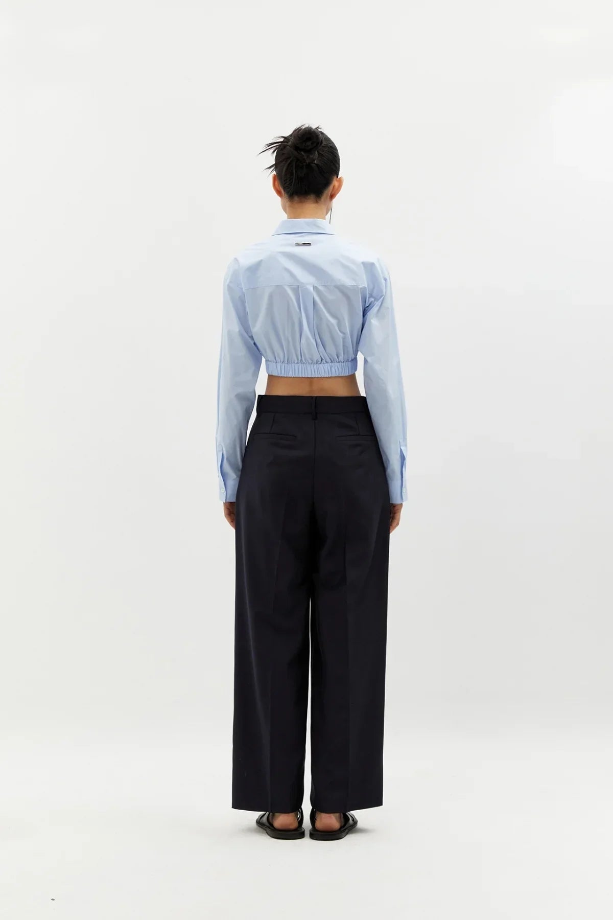 Rear view of woman wearing sky blue cropped cotton shirt and wide-leg black trousers on white background