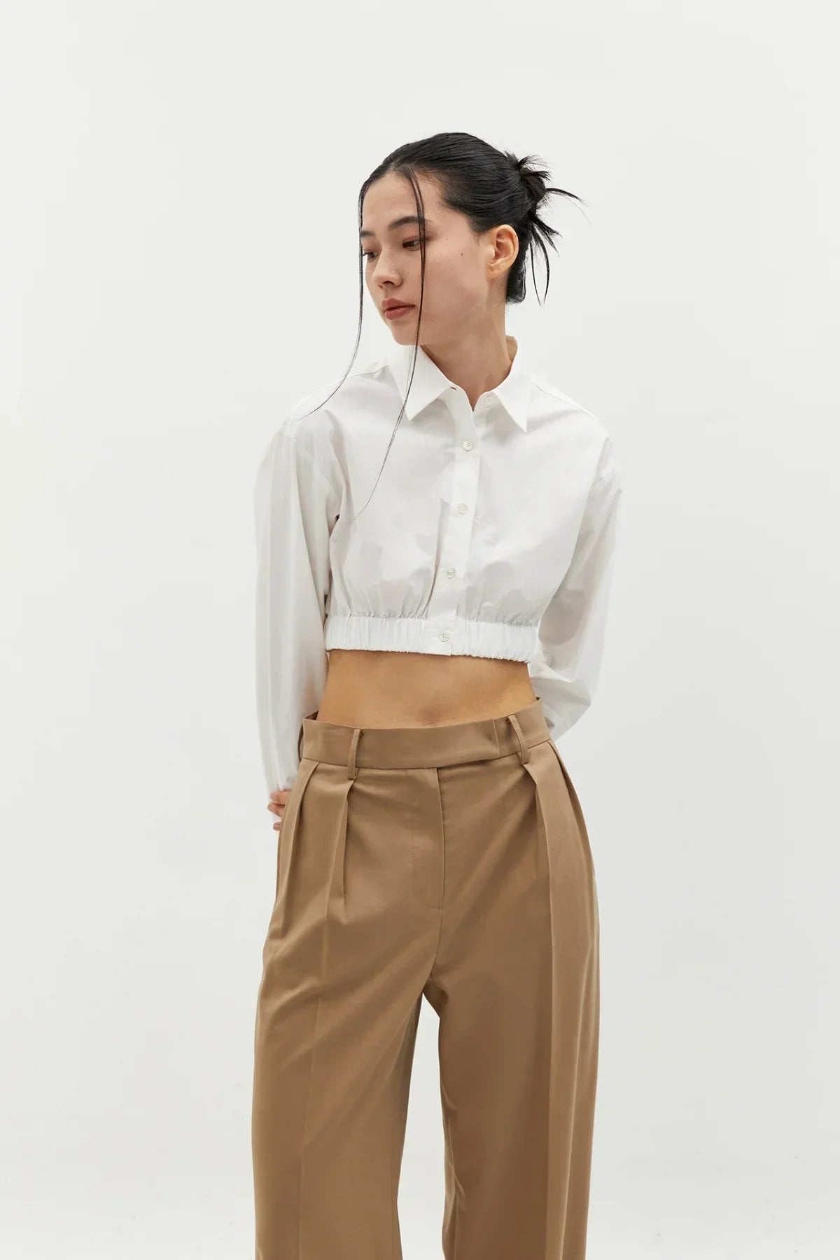 Woman wearing a white cropped cotton shirt and high-waisted beige pleated pants in a studio