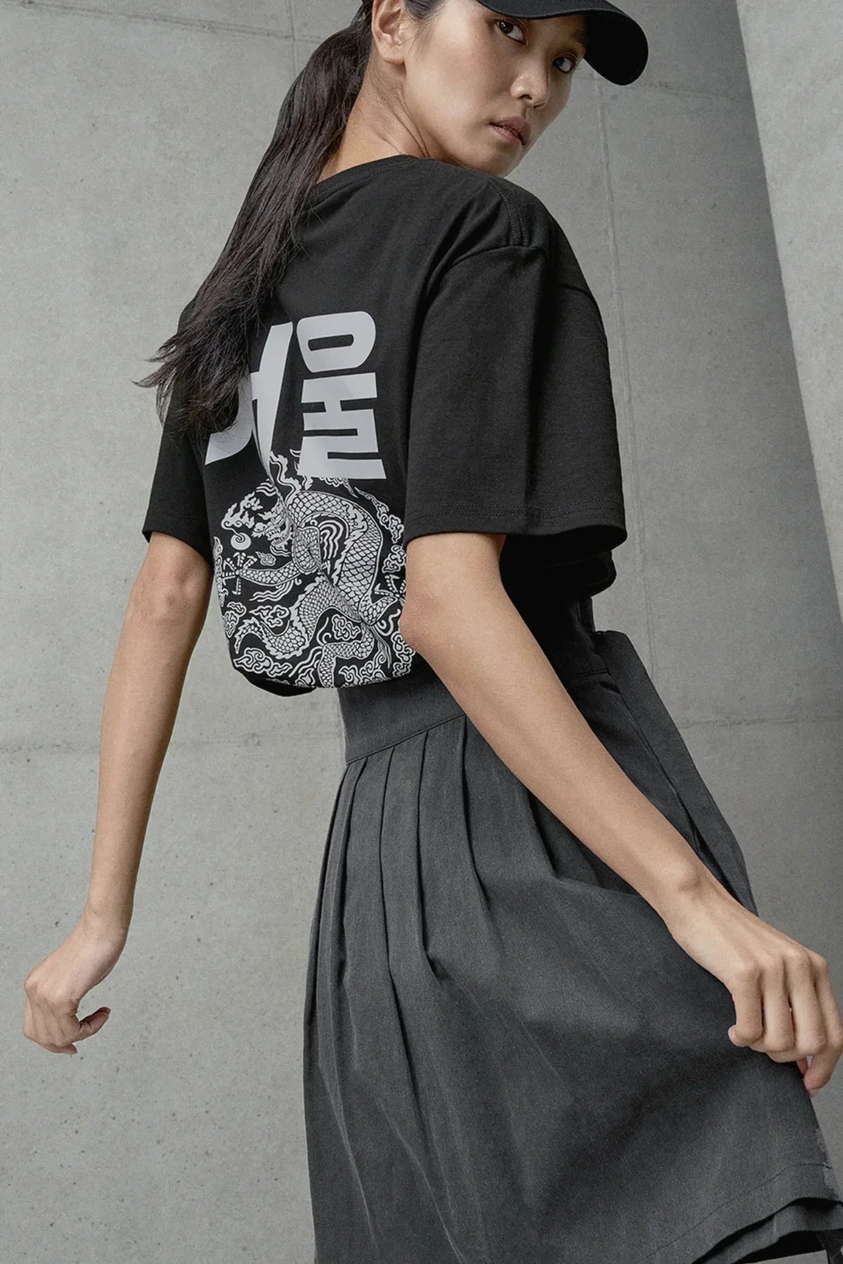 Woman wearing black graphic t-shirt with Korean print and dragon design paired with high-waisted pleated gray skirt