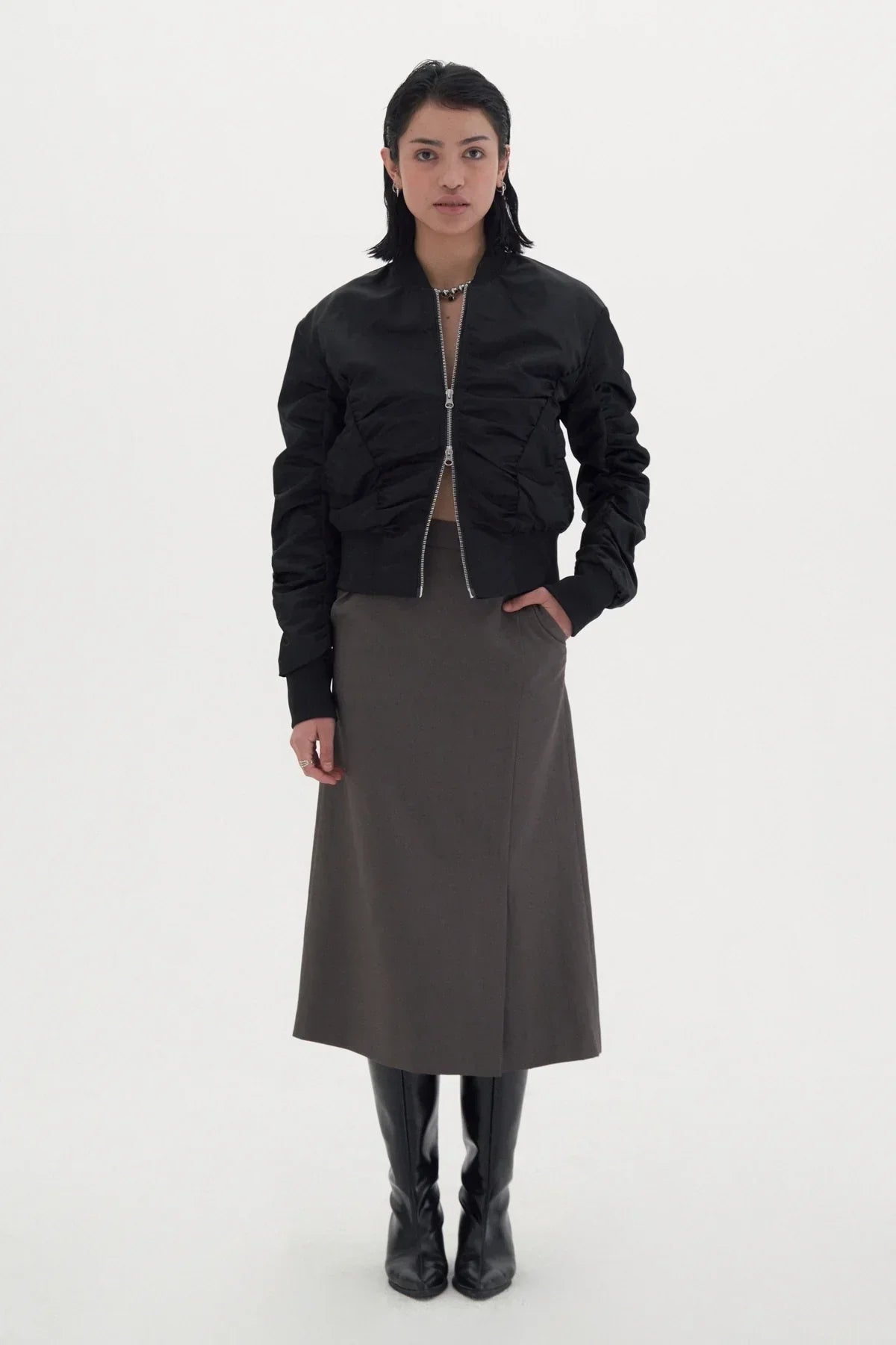 Woman wearing black shirred zip-up jacket, gray midi skirt, and black knee-high boots on white background