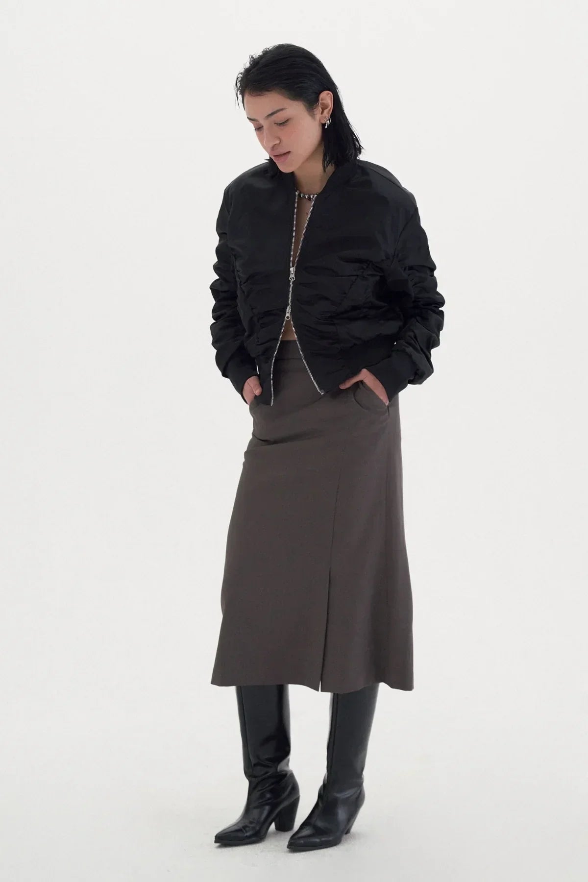 Woman wearing black shirred bomber jacket, brown midi skirt, and black knee-high boots on white background