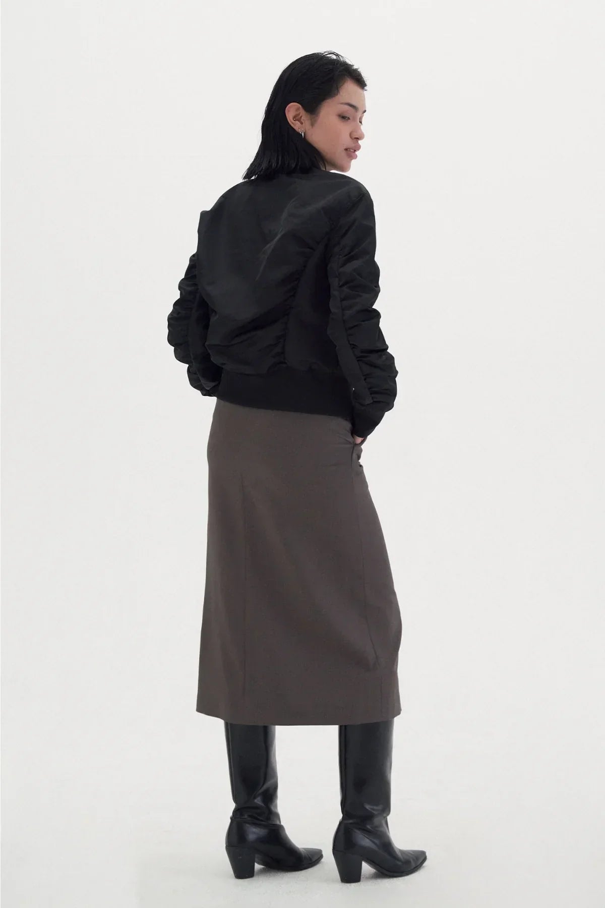Woman wearing black shirred jacket, brown midi skirt, and black knee-high boots on white background