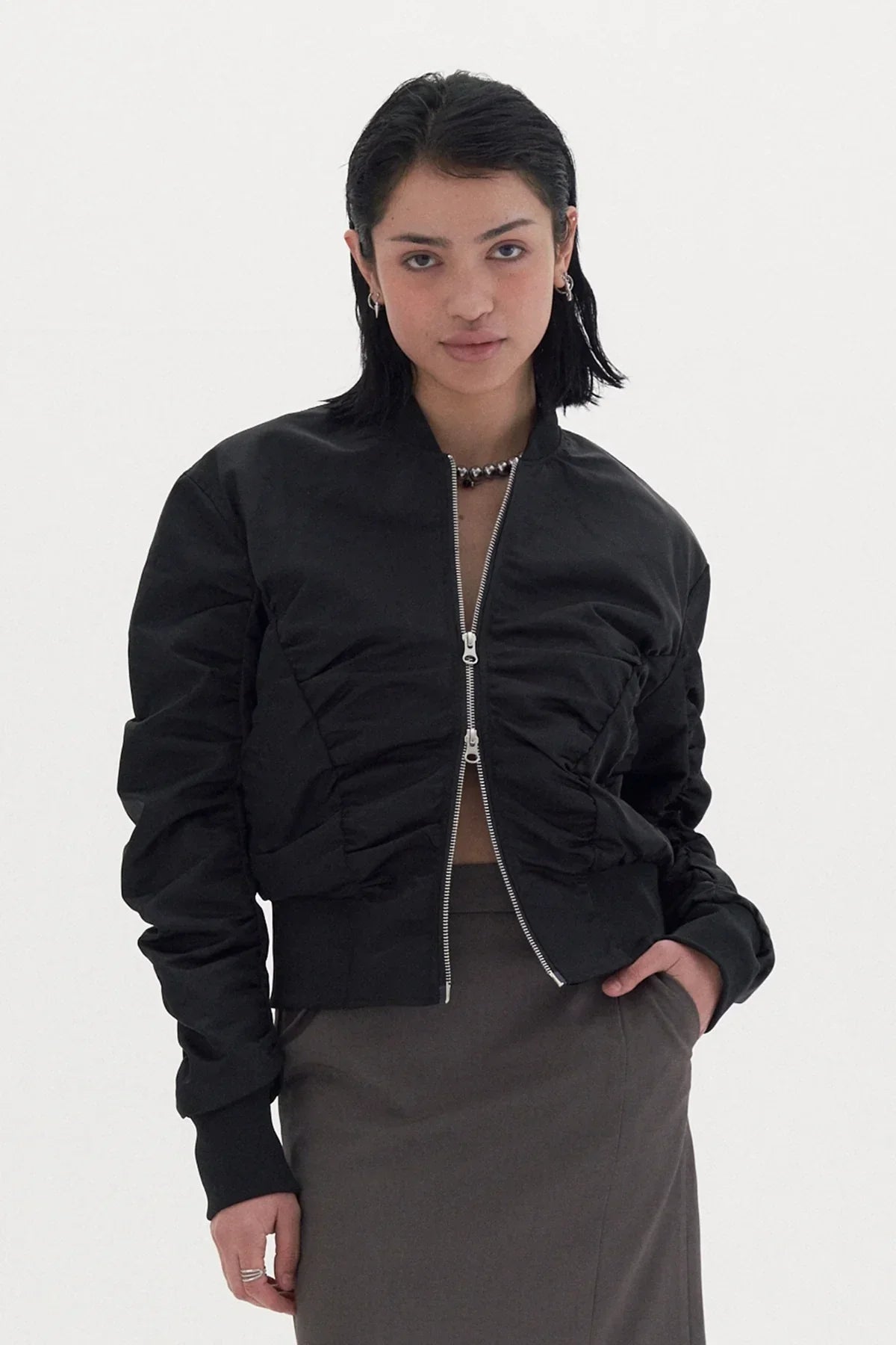 Young woman wearing black shirred bomber jacket with zipper and gray skirt in studio