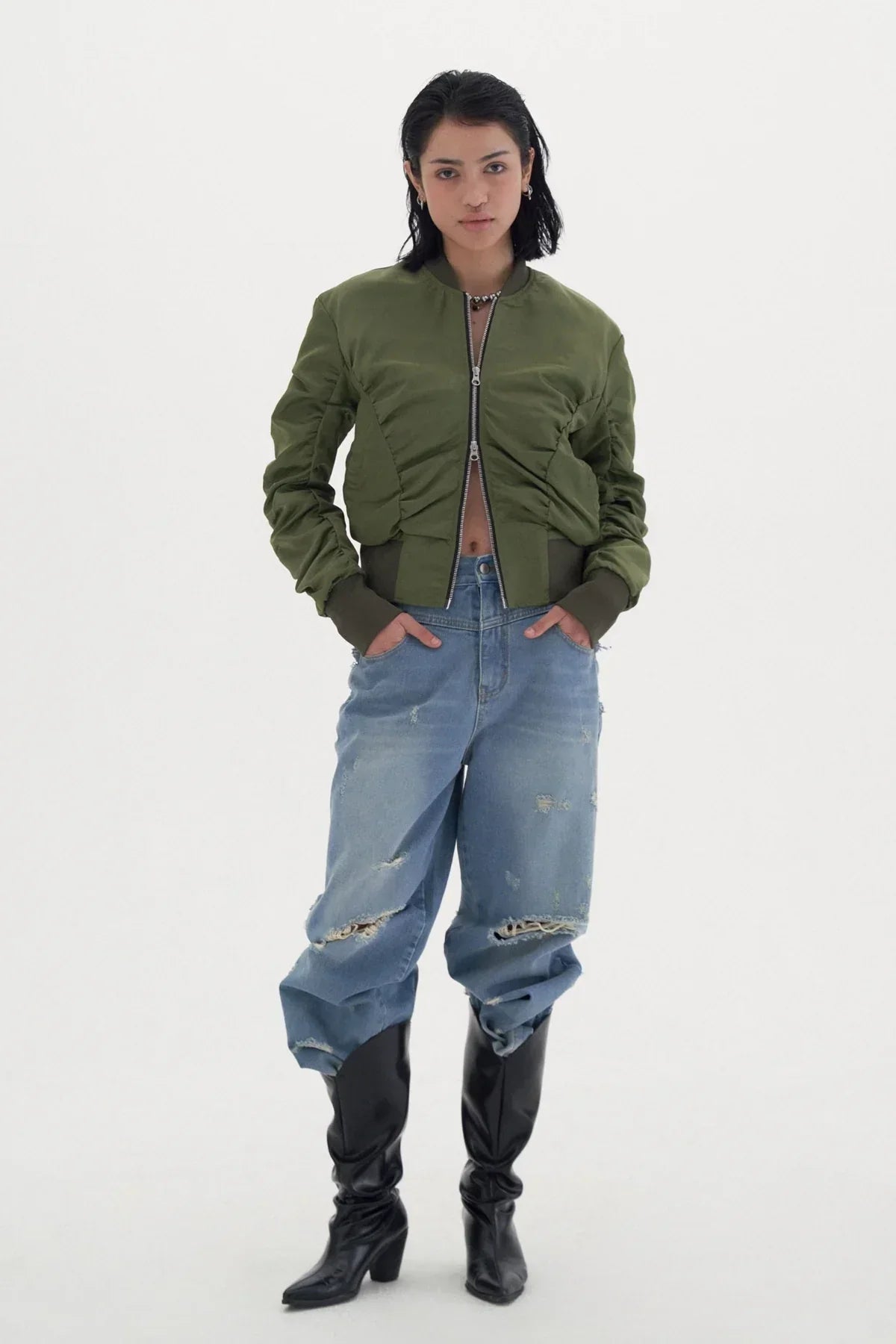 Woman in khaki green shirred bomber jacket, distressed baggy jeans, and black heeled boots on white background