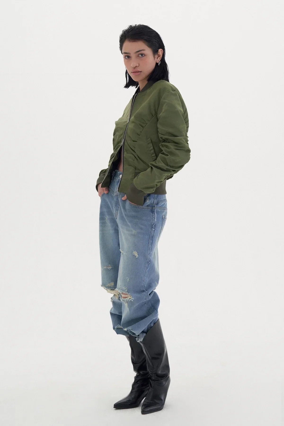 Model wearing olive green bomber jacket, distressed baggy jeans, and black pointed boots on white background