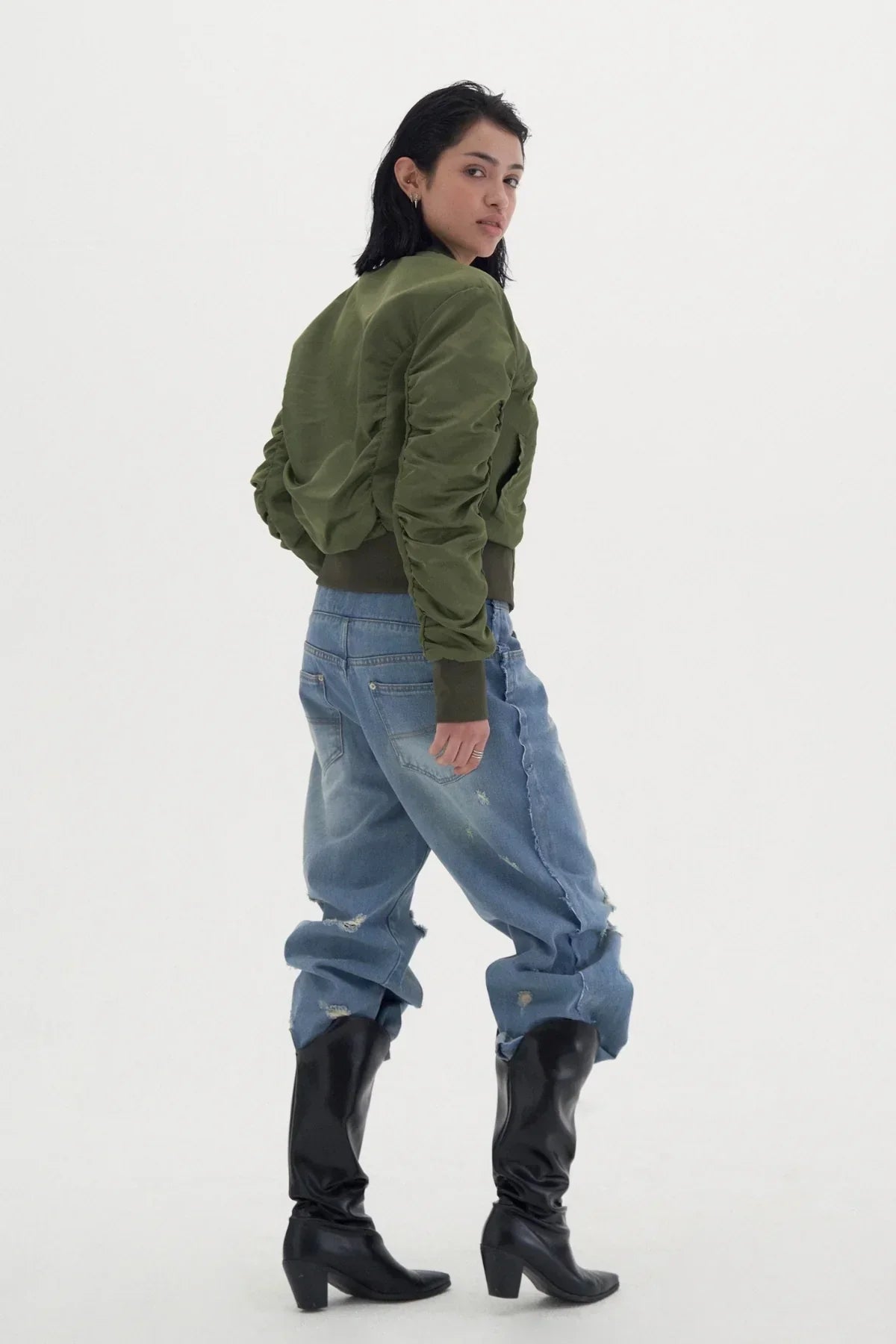 Woman wearing green shirred bomber jacket, distressed baggy jeans, and black heeled boots posing in studio