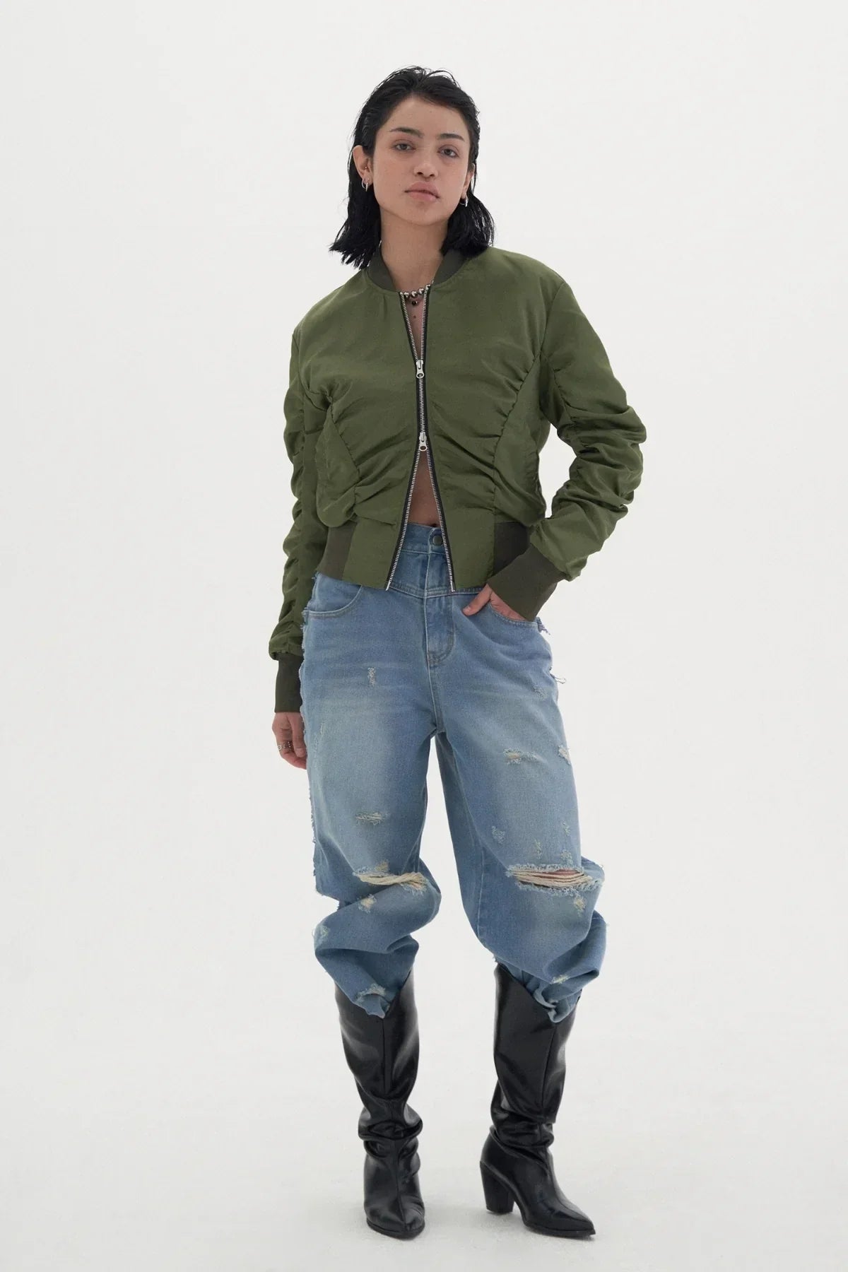 Woman wearing khaki shirred bomber jacket, ripped blue jeans, and black heeled boots on white background
