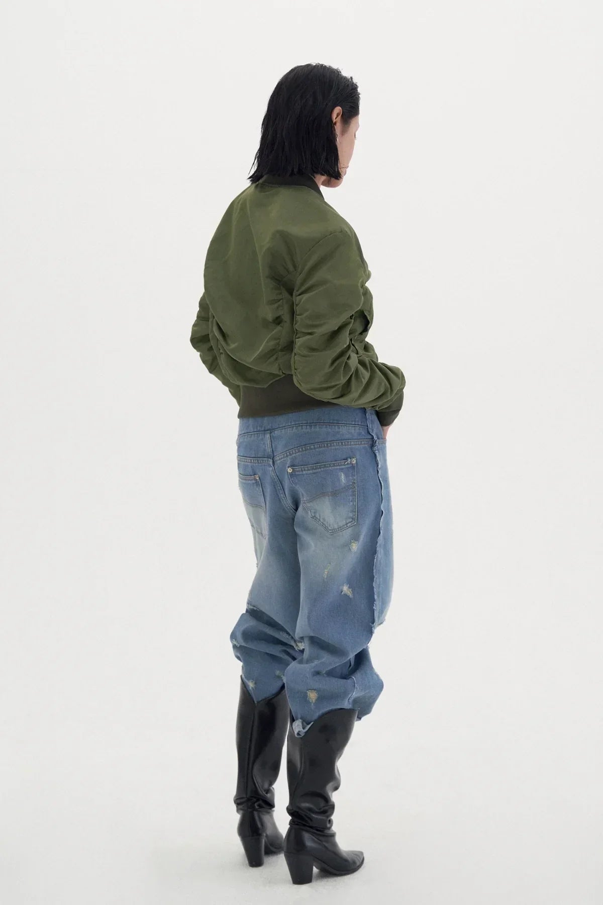 Woman wearing khaki shirred bomber jacket with distressed blue jeans and black heeled boots on white background
