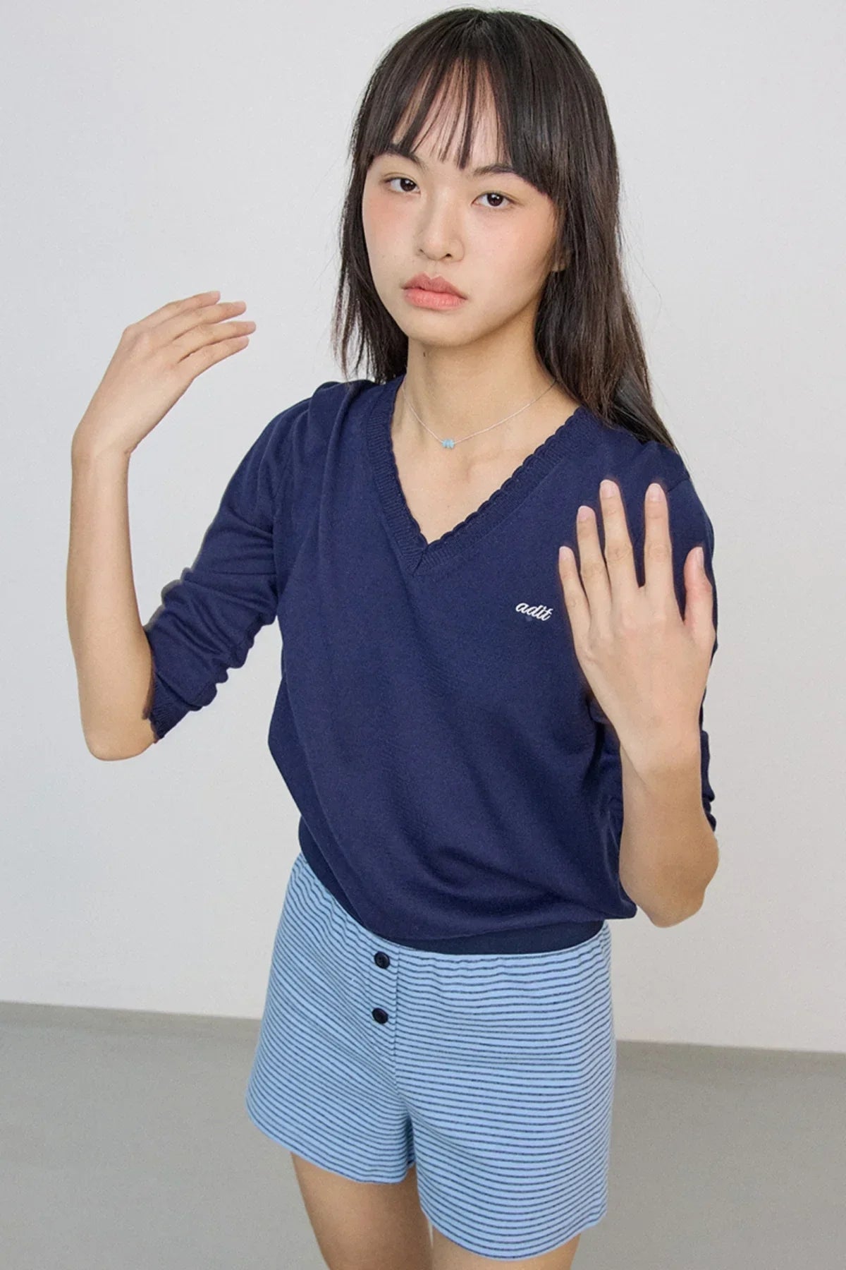 Woman wearing blue small bead necklace, navy sweater, and striped blue shorts against plain background