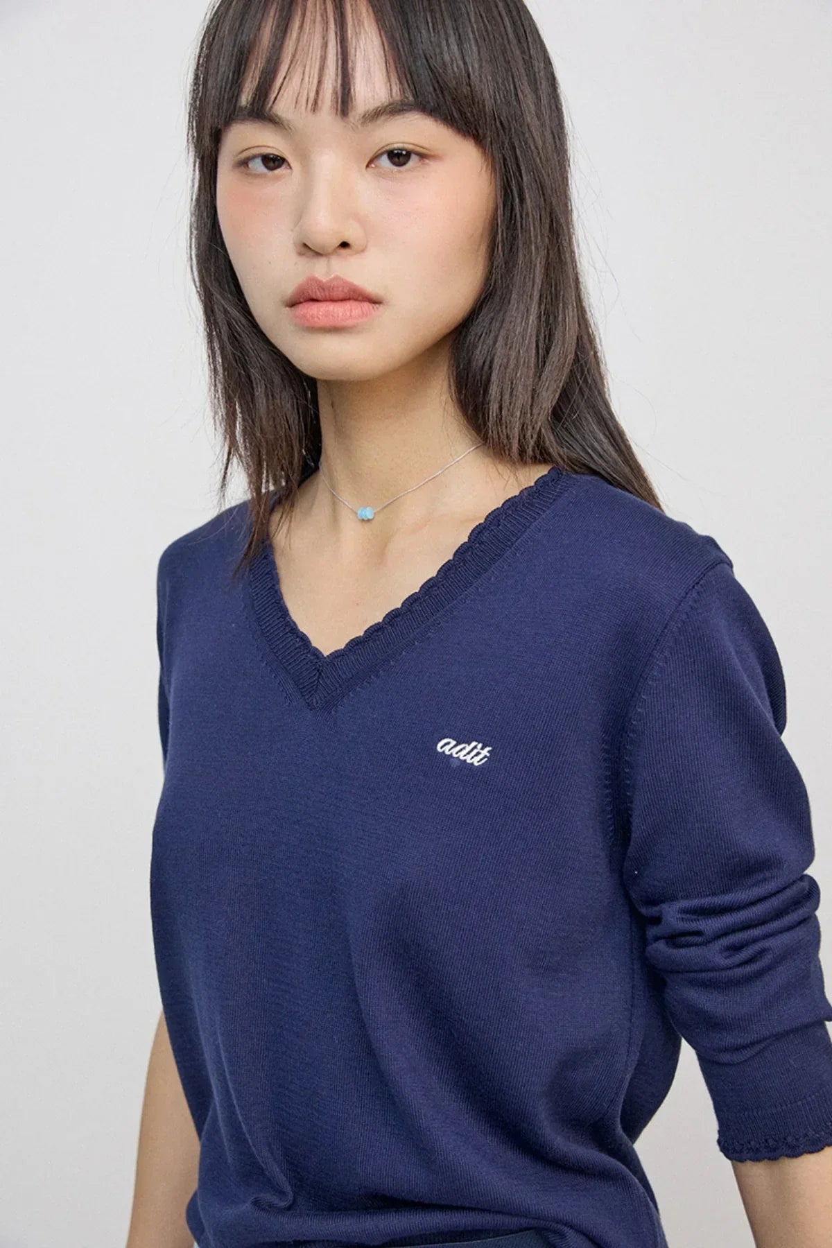 Young woman wearing a blue V-neck sweater with 'adit' embroidery and a delicate blue bead necklace