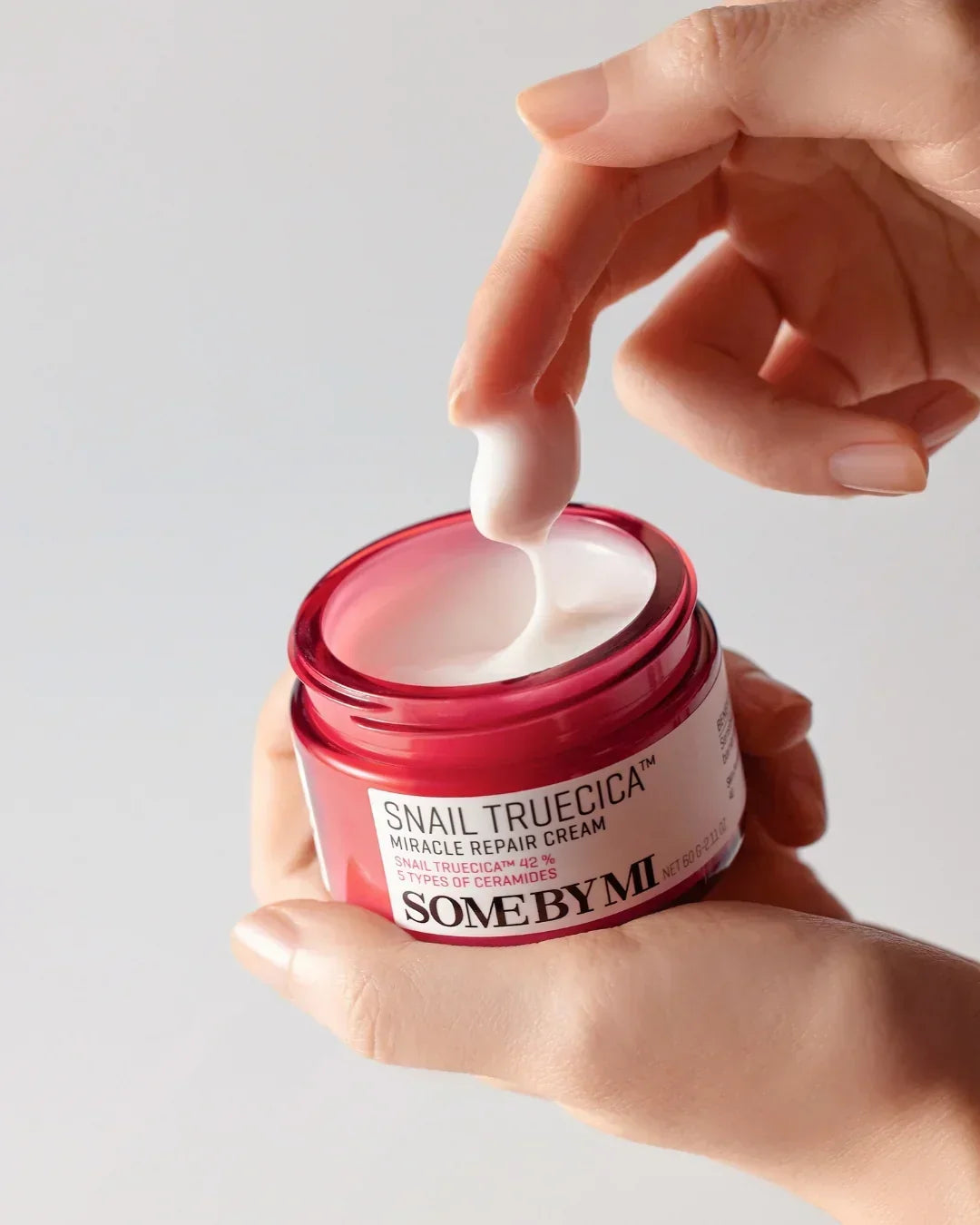 Hand scooping white Snail Truecica Miracle Repair Cream from red jar by Some By Mi