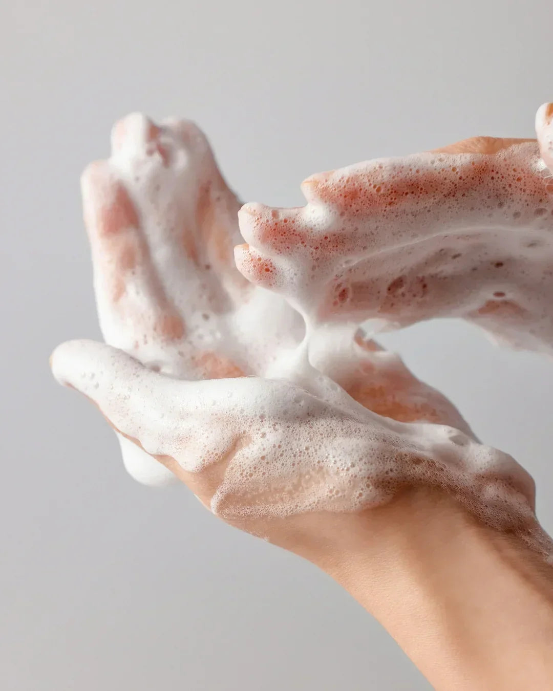Hands covered in thick white foam against a gray background, demonstrating cleansing or skincare