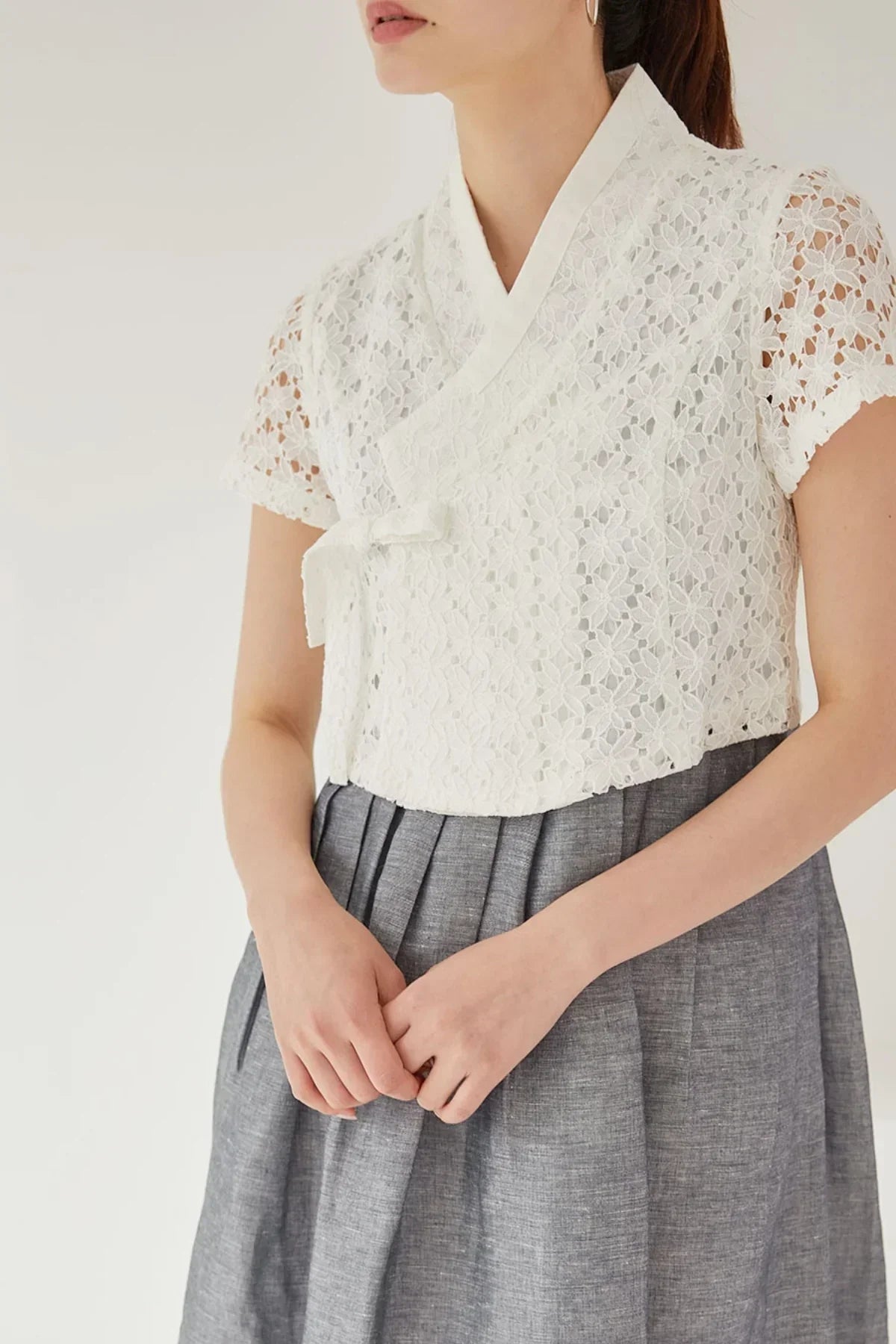 Woman wearing white floral lace jeogori top paired with gray pleated skirt, standing with hands clasped