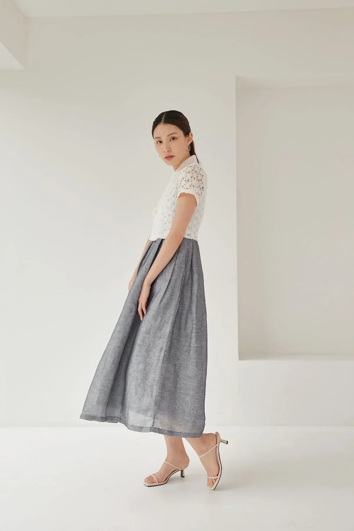 Asian woman in white lace blouse and gray midi skirt posing in minimalist white studio with beige heels