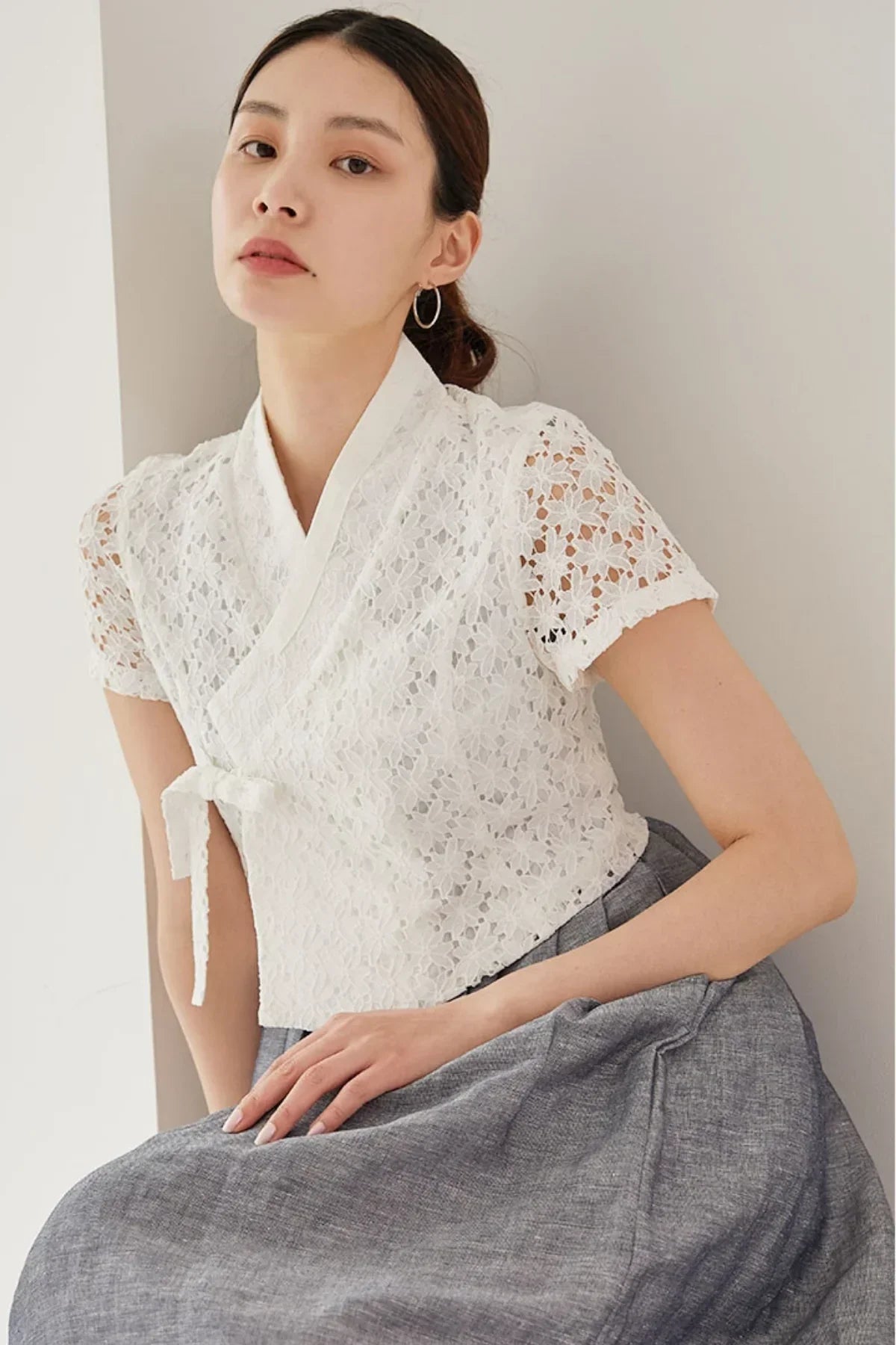 Asian woman wearing white floral lace short-sleeve jeogori top and gray skirt leaning on wall