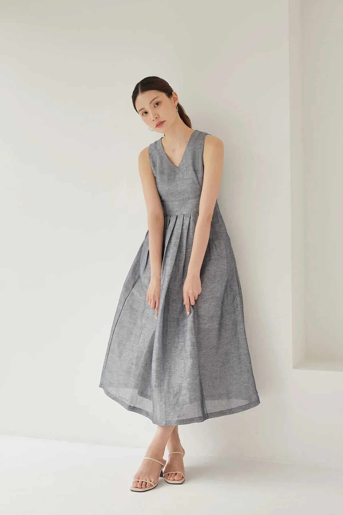 Woman in sleeveless gray midi dress and beige heels posing against white wall in minimalist studio