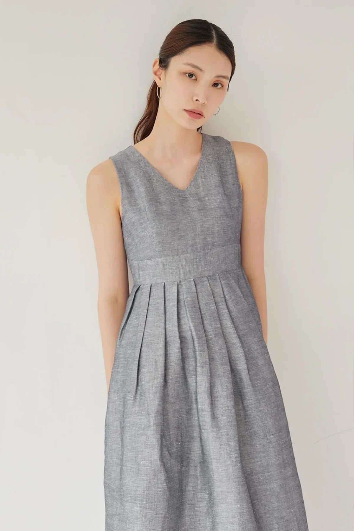 Young woman wearing sleeveless gray linen dress with pleated skirt standing against white wall