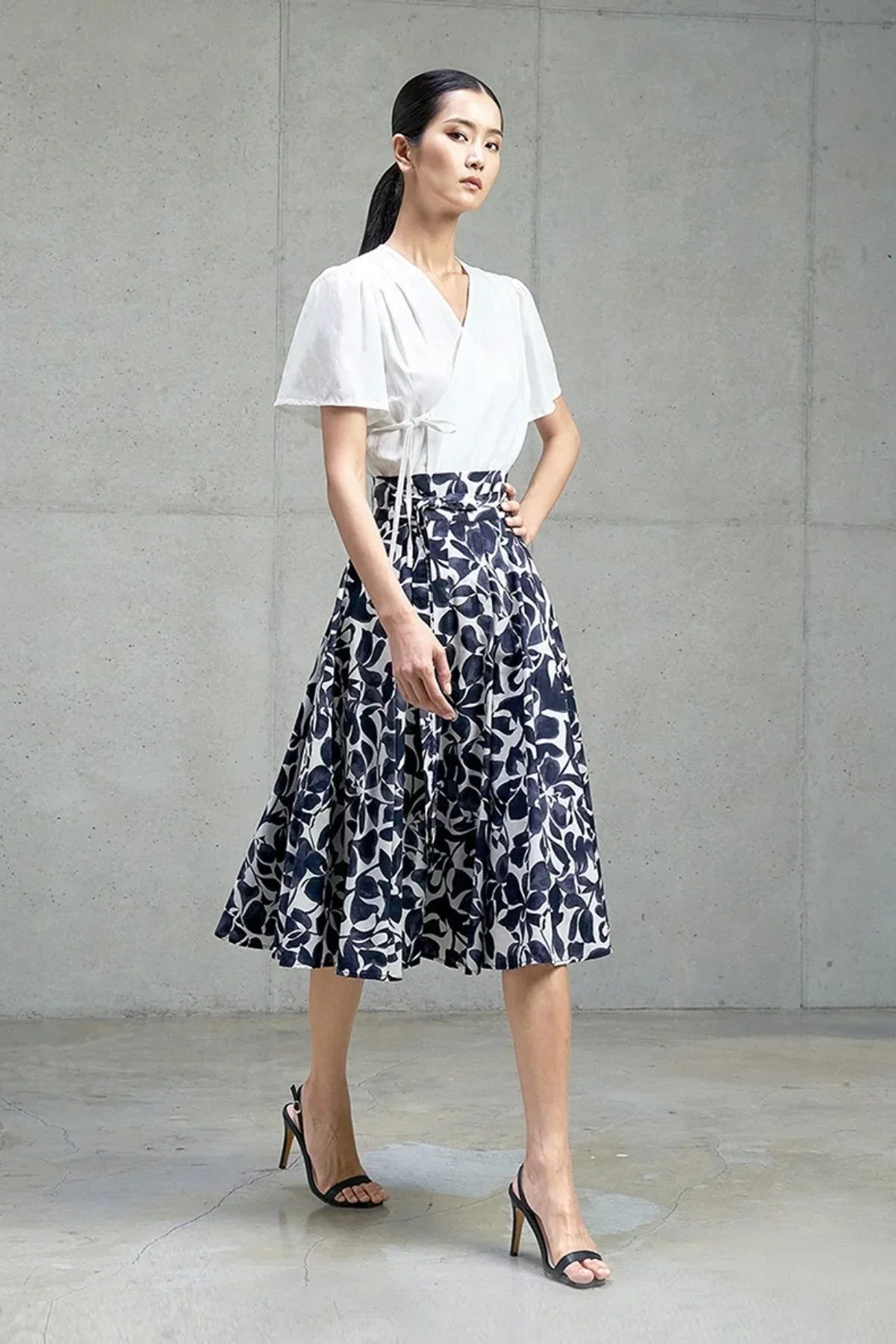 Model wearing a white wrap top paired with a navy floral print midi skirt and black high heel sandals in a minimalist studio