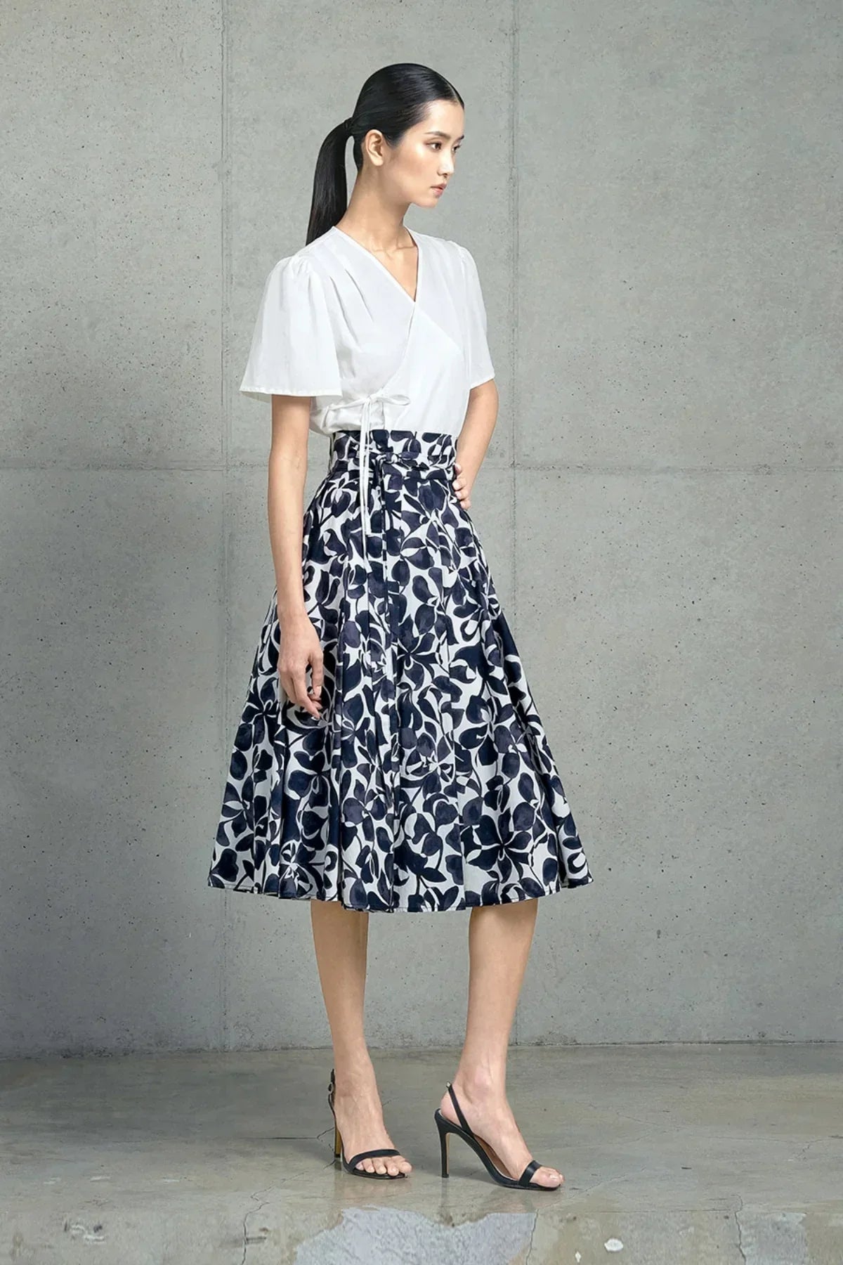 Woman wearing white wrap blouse and navy floral midi skirt with high heels against gray concrete wall