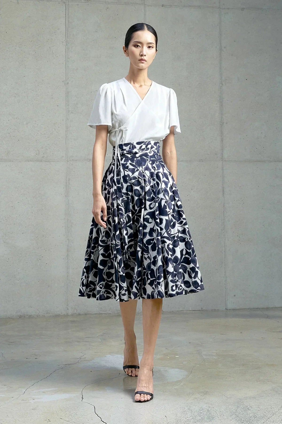 Model wearing a white wrap blouse and navy floral pleated midi skirt in a minimalist studio