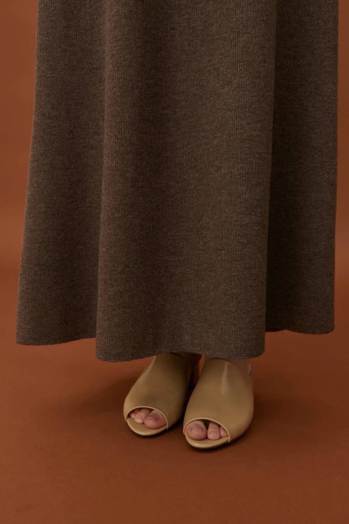 Close-up of brown knit A-line skirt with beige open-toe mule shoes on brown backdrop
