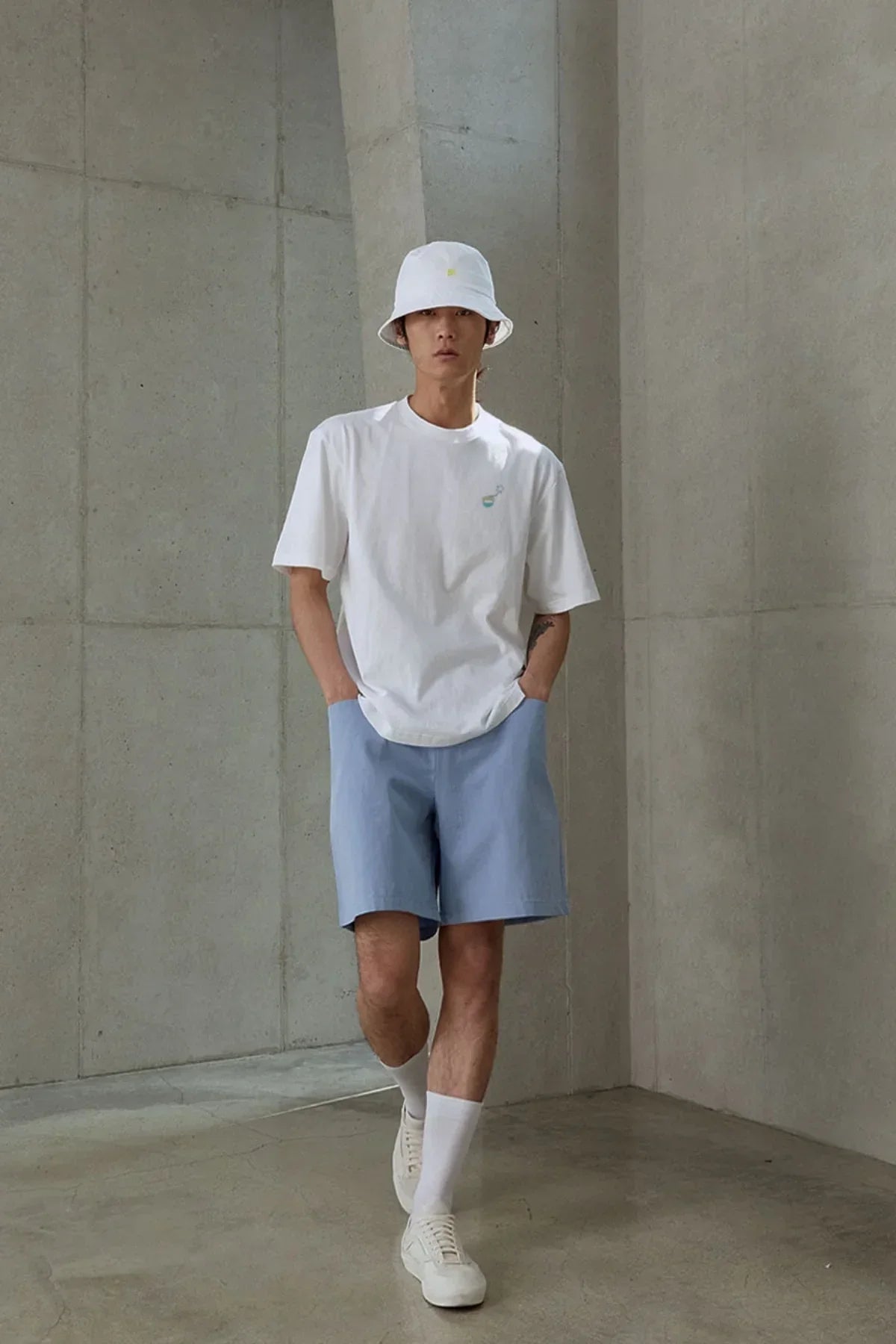Man wearing white graphic t-shirt, white bucket hat, light blue shorts, white socks, and white sneakers in minimalist concrete room