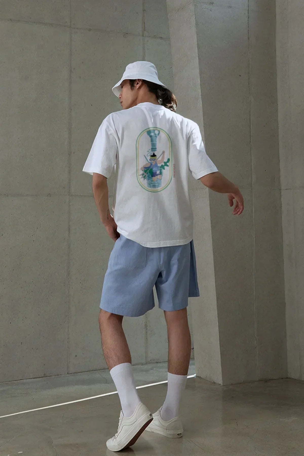 Man wearing white graphic t-shirt, light blue shorts, white bucket hat, and white sneakers in minimal concrete setting