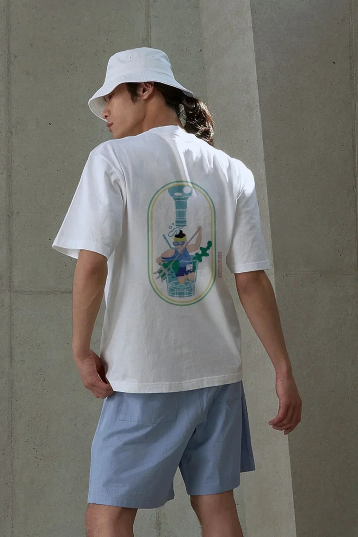 Man wearing white graphic t-shirt with colorful scuba diving print and light blue shorts against gray concrete wall