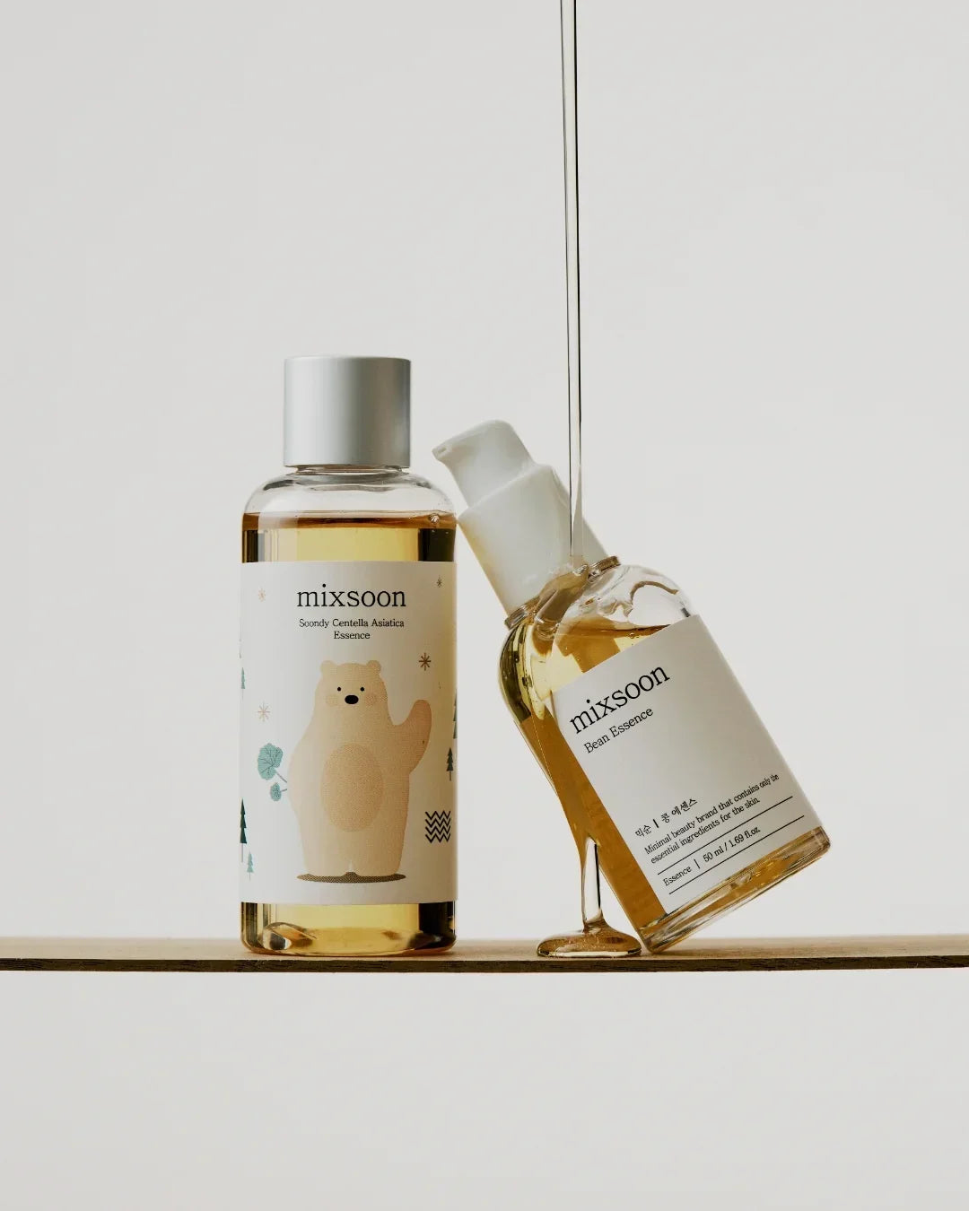 Two mixsoon skincare essence bottles on a wooden surface with pouring liquid, minimalist background