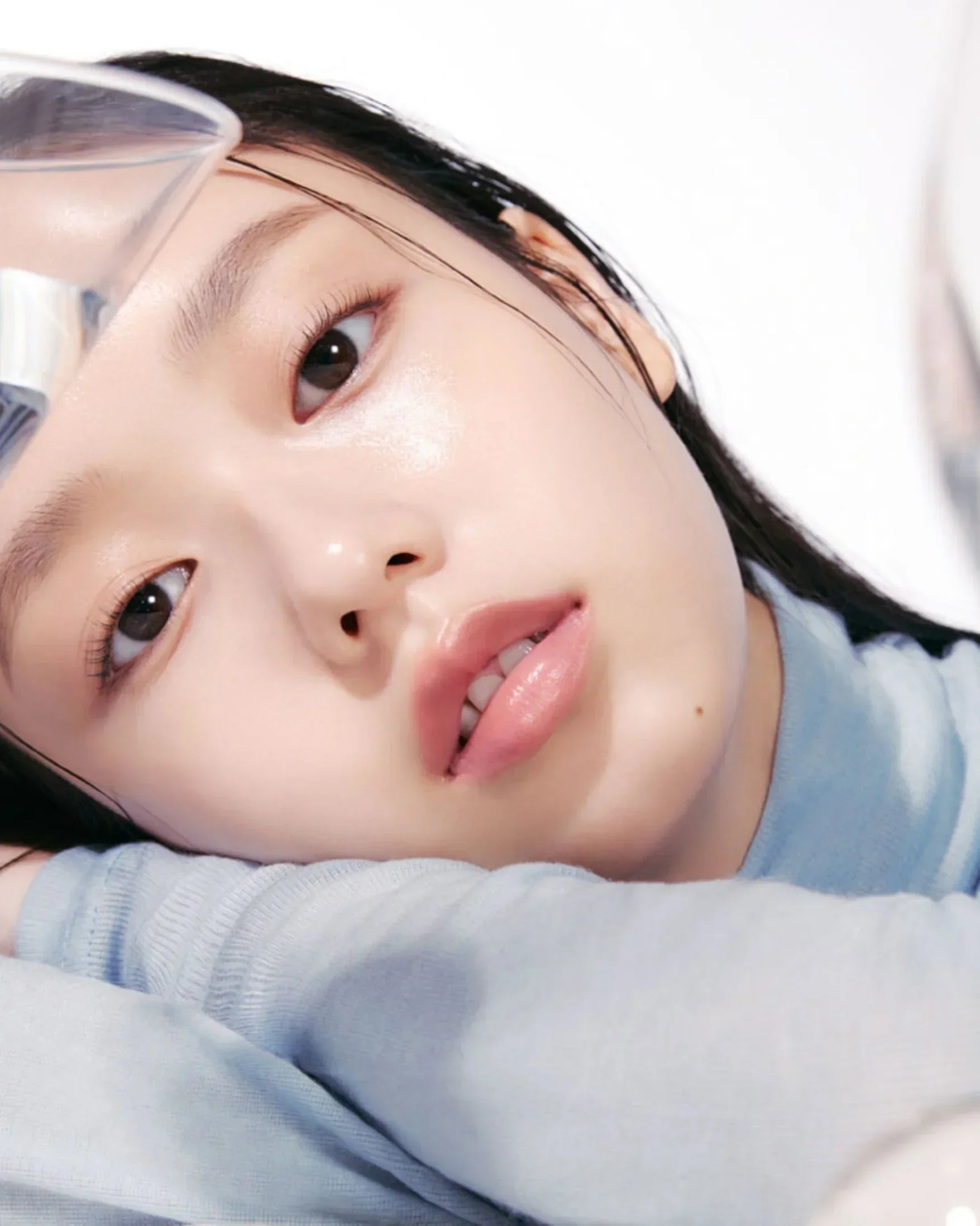 Close-up of a young woman with flawless glowing skin resting on her arm, wearing a light blue top