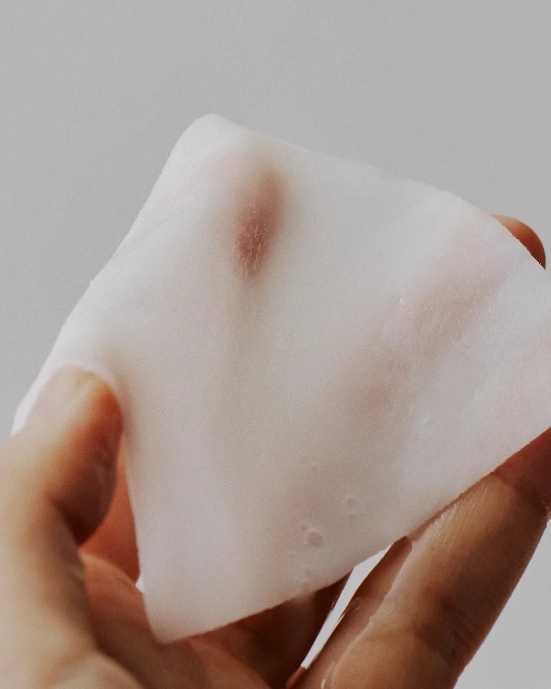 Hand holding a translucent white facial cotton pad against a plain gray background