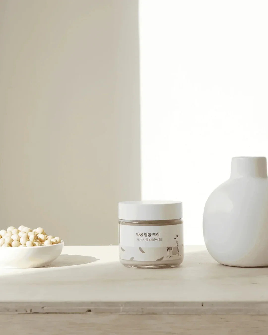 Soybean nourishing cream jar with Korean label on white surface, next to white bowl of soybeans and white vase