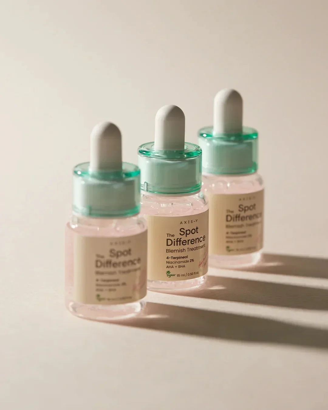 Three bottles of Axis-Y The Spot Difference Blemish Treatment with dropper caps on a beige surface