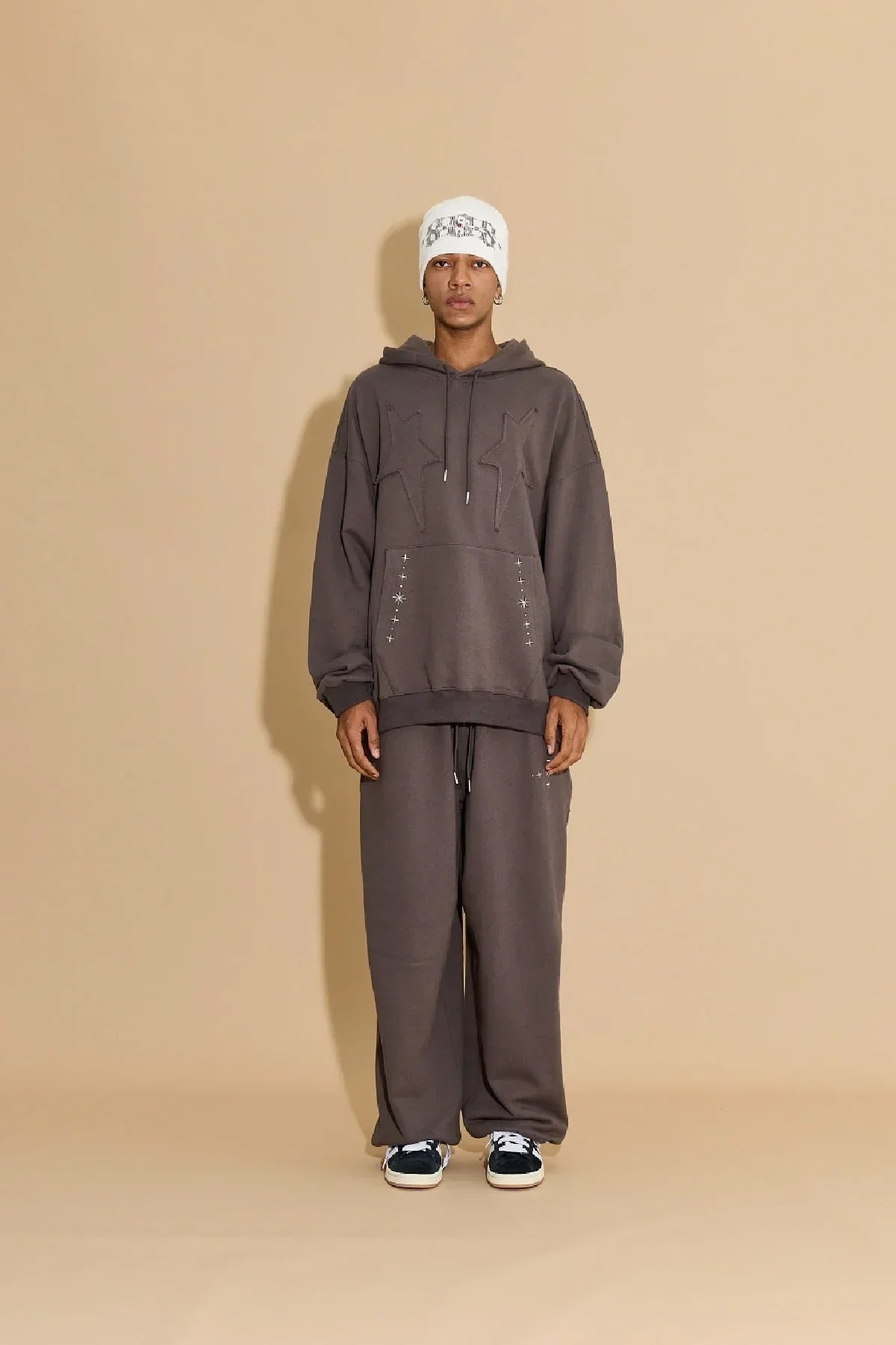 Model wearing charcoal oversized hoodie and pants with star embroidery and white beanie on beige background