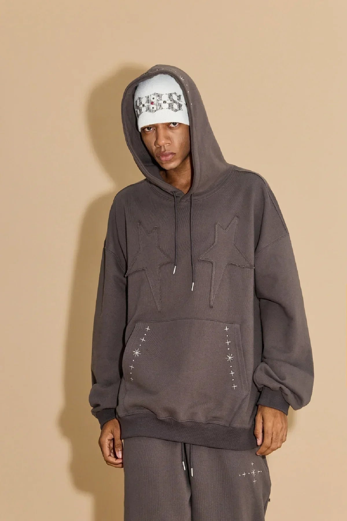 Model wearing charcoal hoodie with star applique embroidery and matching sweatpants against beige background