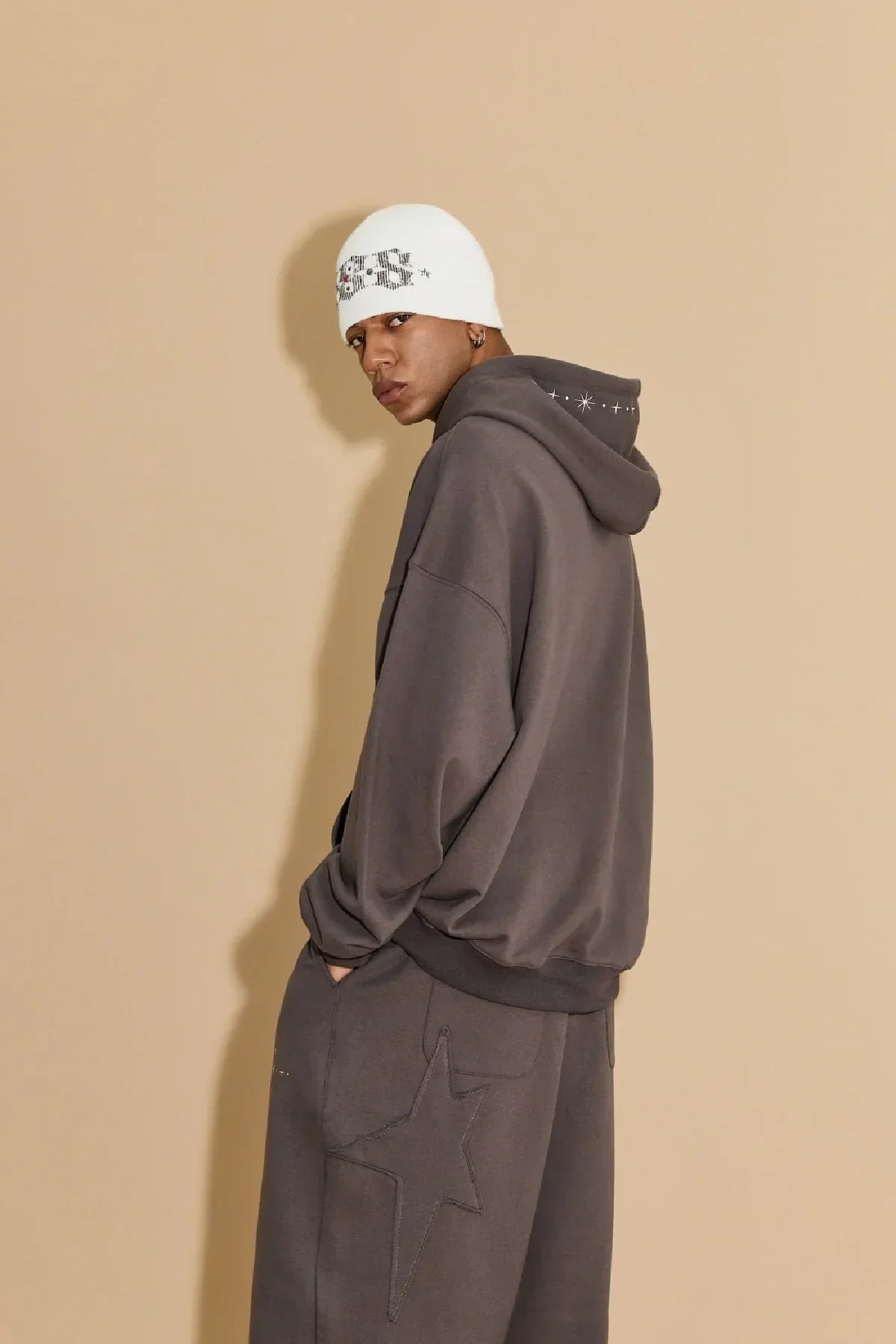 Model wearing charcoal star appliqué hoodie and pants with white beanie against beige background
