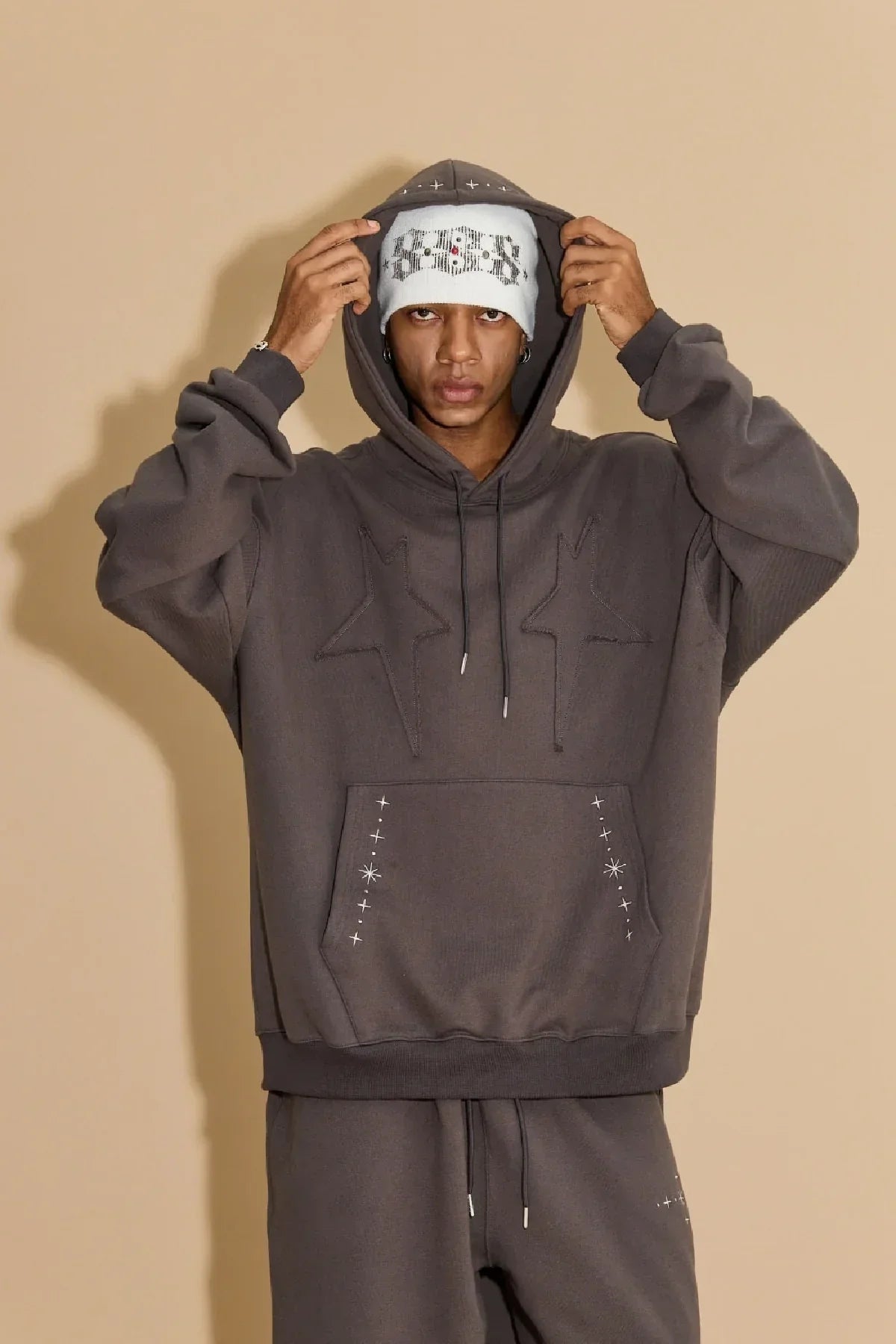 Man wearing charcoal hoodie with embroidered stars and matching sweatpants against beige background