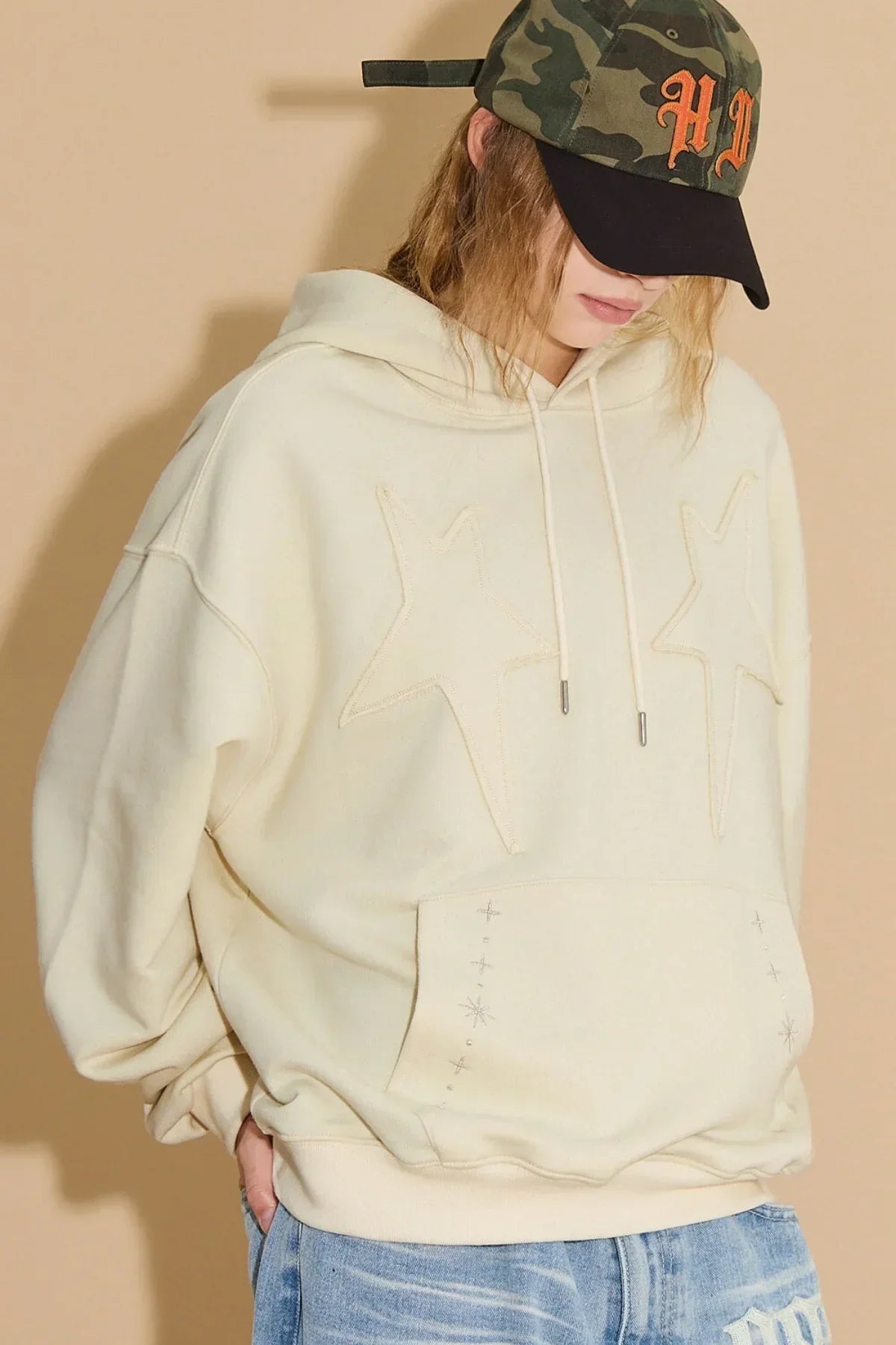 Close-up of person wearing ivory hoodie with star appliques and front pocket embroidery, paired with camouflage cap