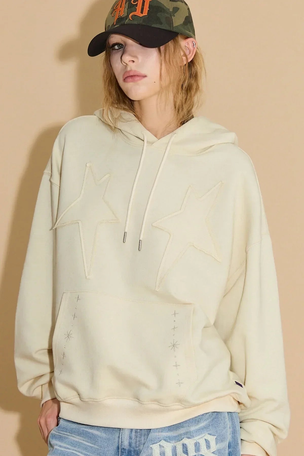Woman wearing ivory hoodie with star appliqué embroidery and light blue jeans against beige background