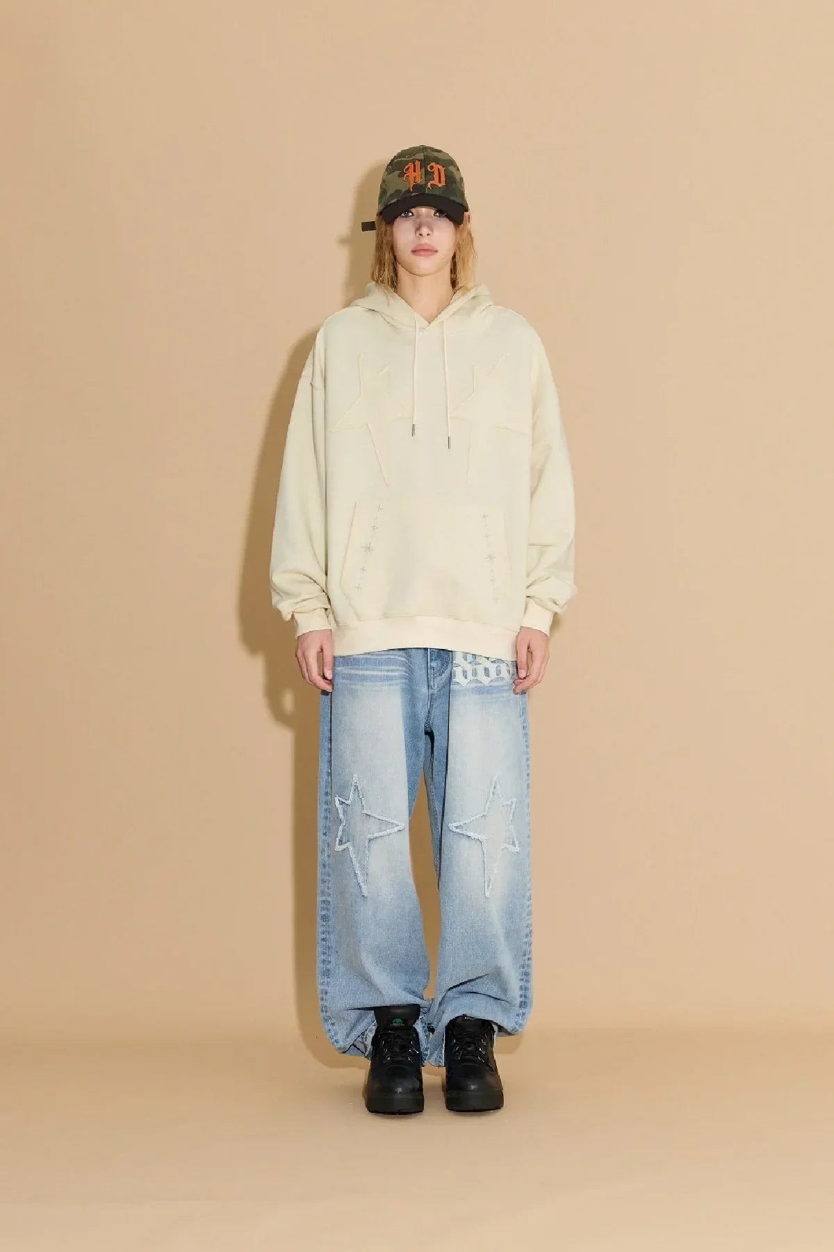 Person wearing ivory star applique hoodie, wide light blue jeans with star patches, black shoes, camo cap