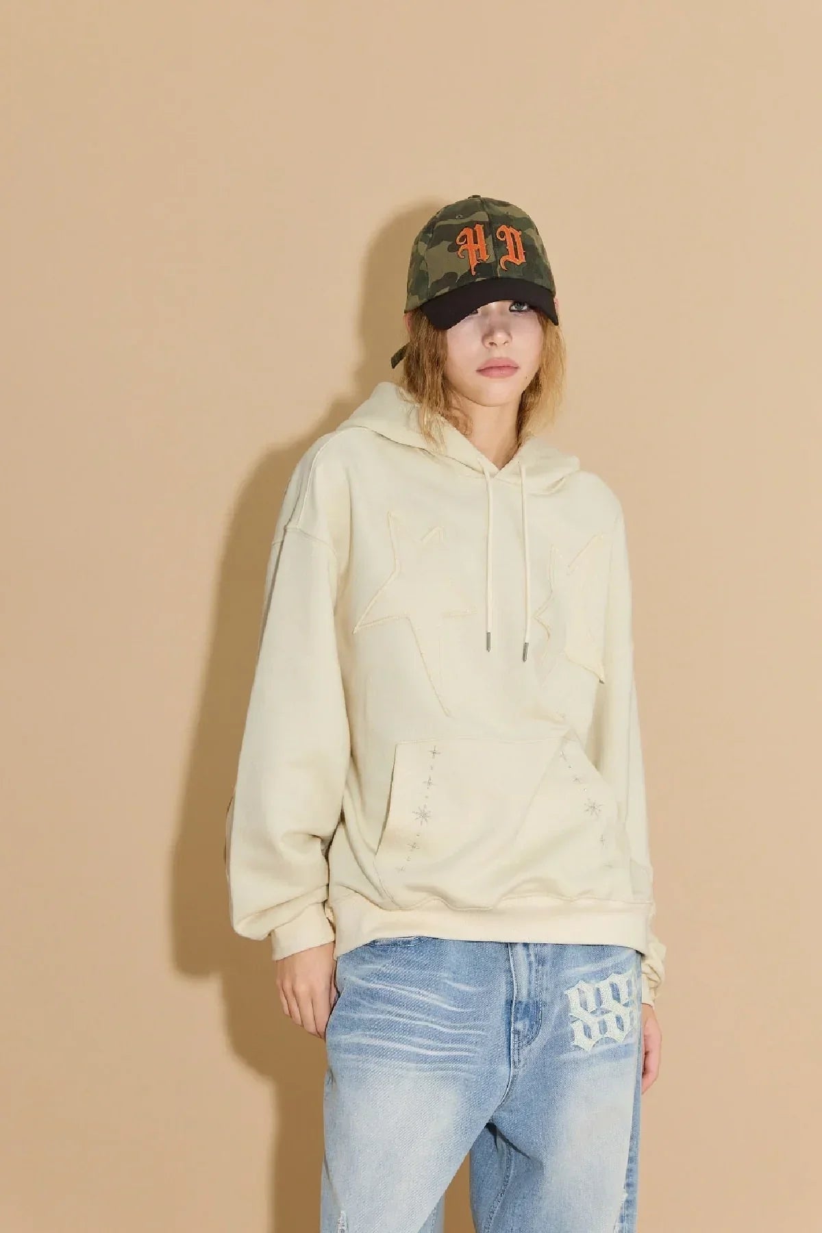 Model wearing ivory star applique embroidery hoodie and blue jeans against beige wall