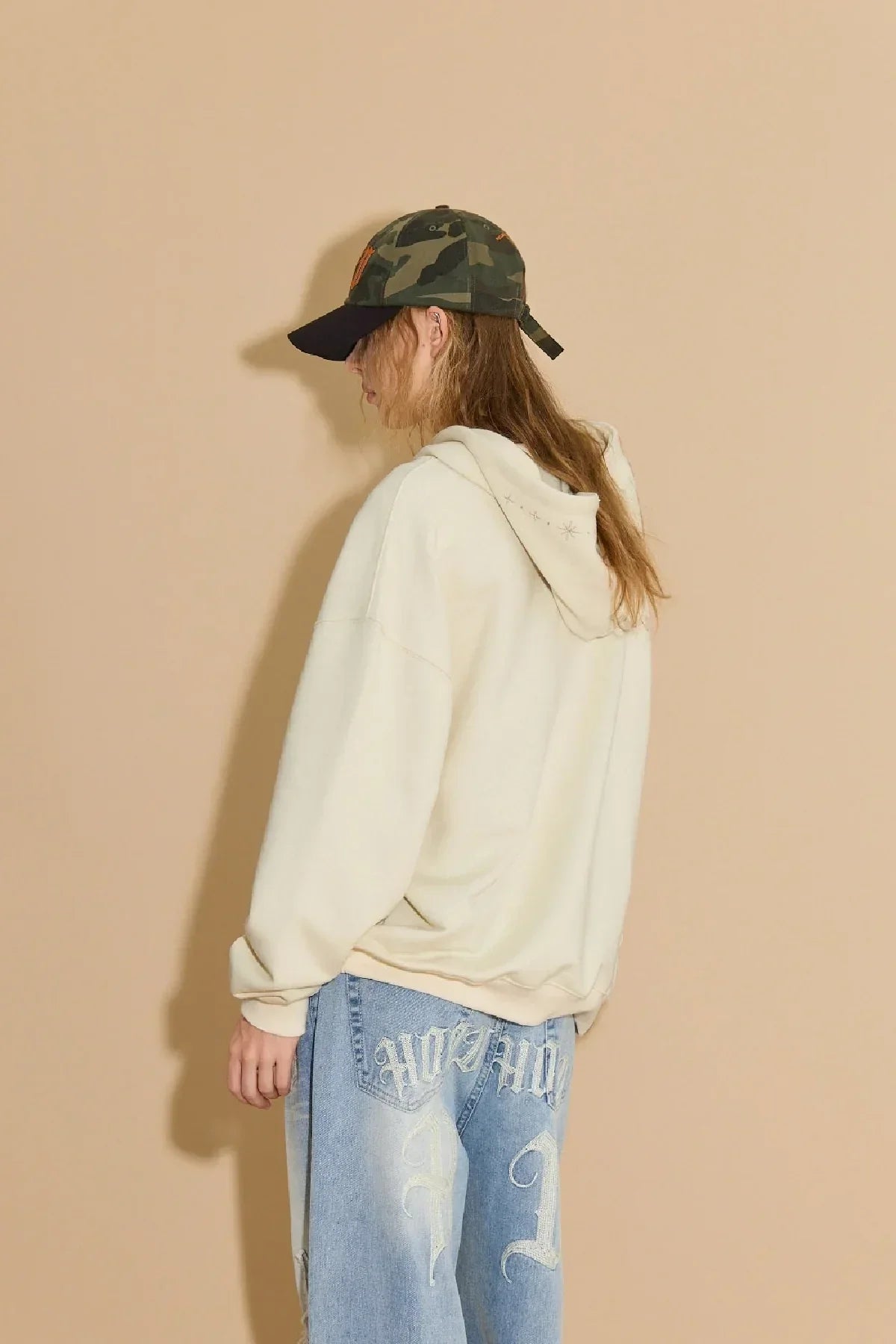 Model in ivory hoodie with star embroidery on hood wearing camo baseball cap and blue jeans with embroidered back pockets